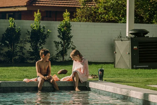 Two children sitting on the edge of a pool with feet in the water near a black generator cover on the lawn.