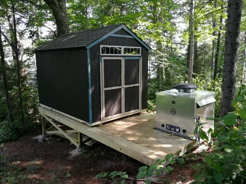 A black shed and silver generator enclosure standing on a wooden platform surrounded by tall trees and green plants.
