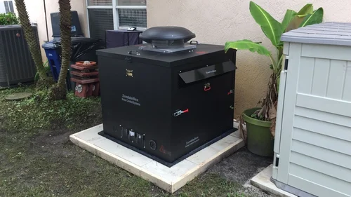 A black outdoor generator box standing on a concrete pad beside a white shed, potted plants, and yard equipment.