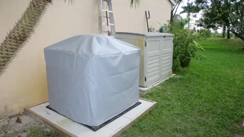 Beside a tan wall, a gray covered generator unit rests on a raised pad near a ladder, besides grass and trees.