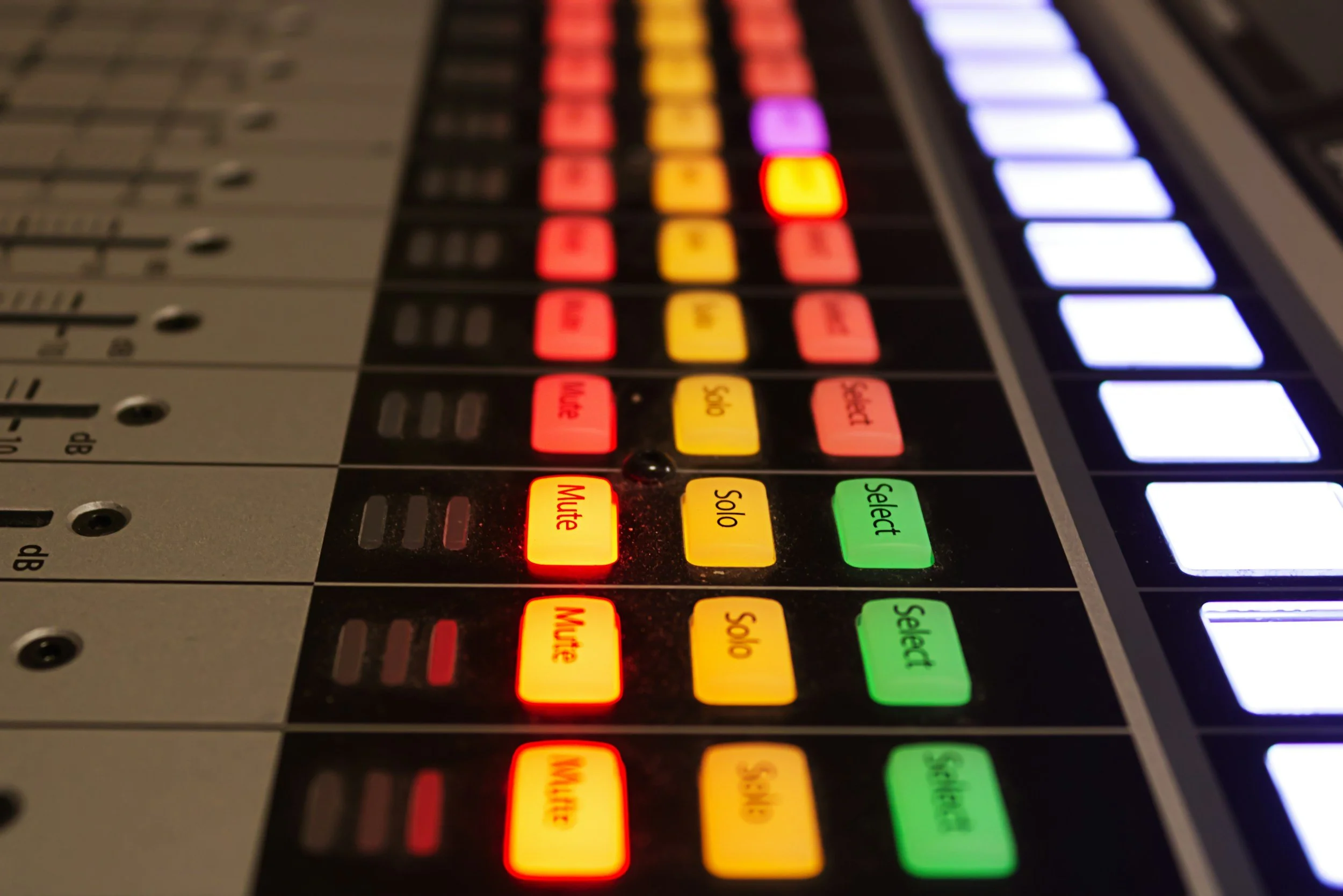 Close-up of a digital audio mixing console with illuminated buttons in red, yellow, green, purple, and white, some labeled with functions such as "Mute" and "Solo."