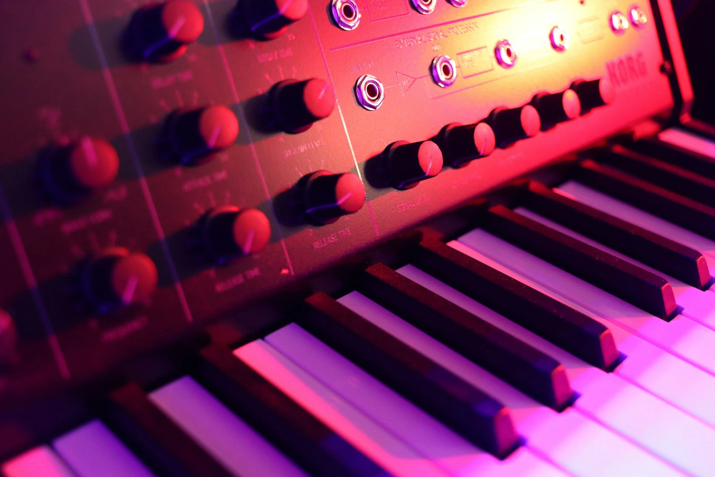 Close-up of a vintage synthesizer keyboard with purple and pink lighting, showing various knobs, buttons, and piano keys.