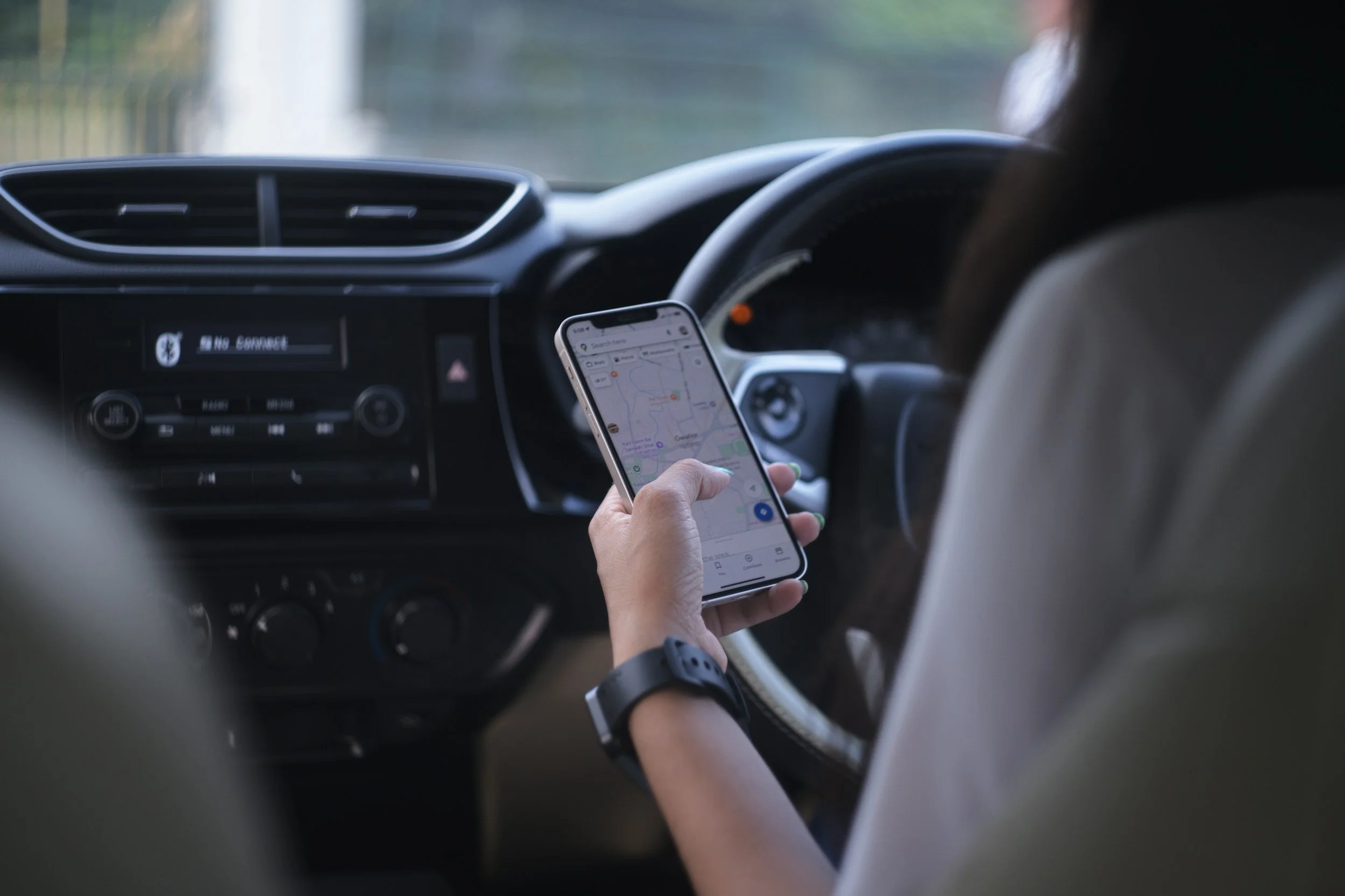 Inside a car with a person holding a smartphone displaying a map, focused on driving, with dashboard controls and an air vent visible.