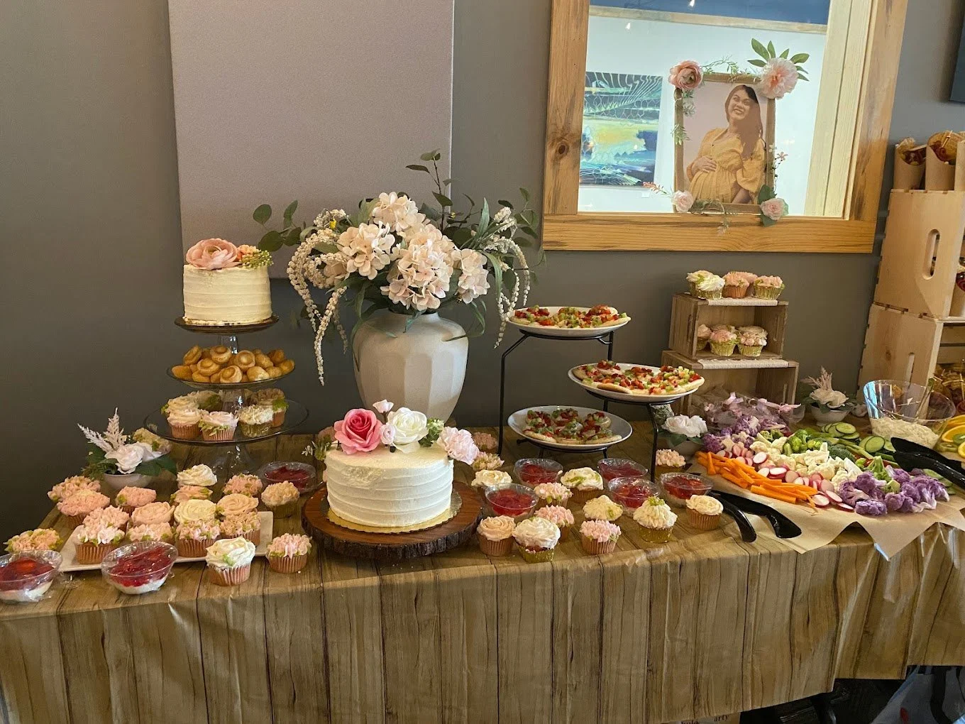 Dessert and snack table with cakes, cupcakes, flowers, and fresh vegetables.