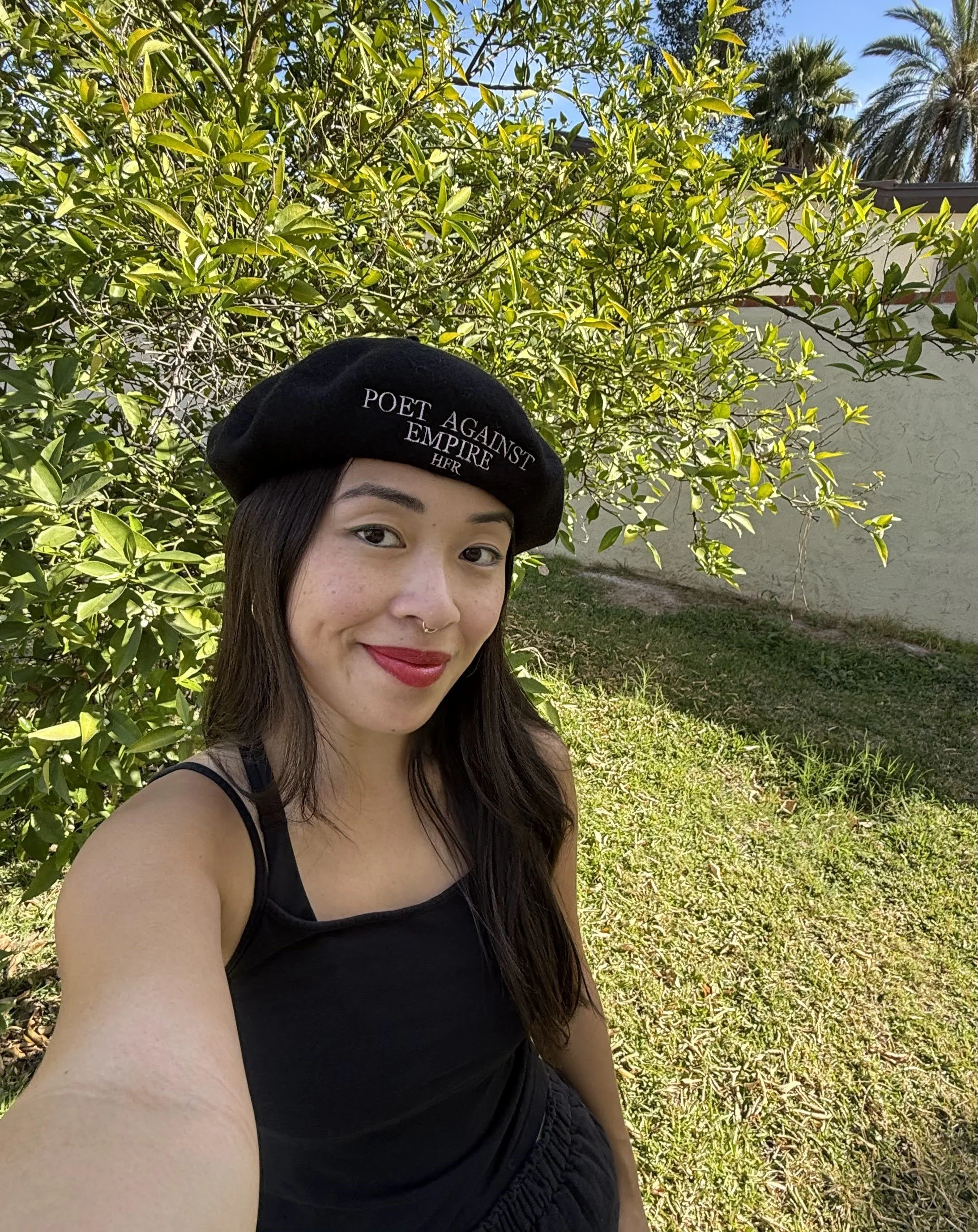 Susan wears black beret and poses in front of green tree and grass.