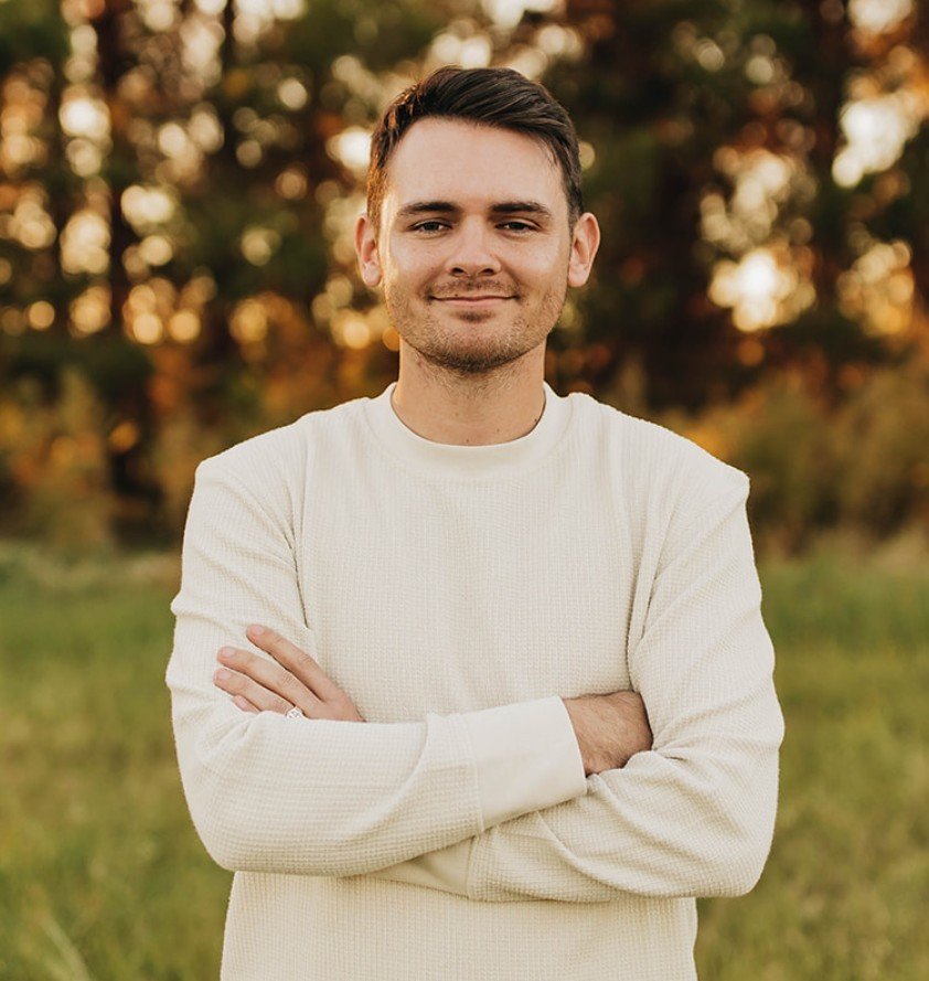 Photo of Cade Anderson from the waist up, wearing a white long-sleeve shirt, and standing in front of an outdoor landscape.