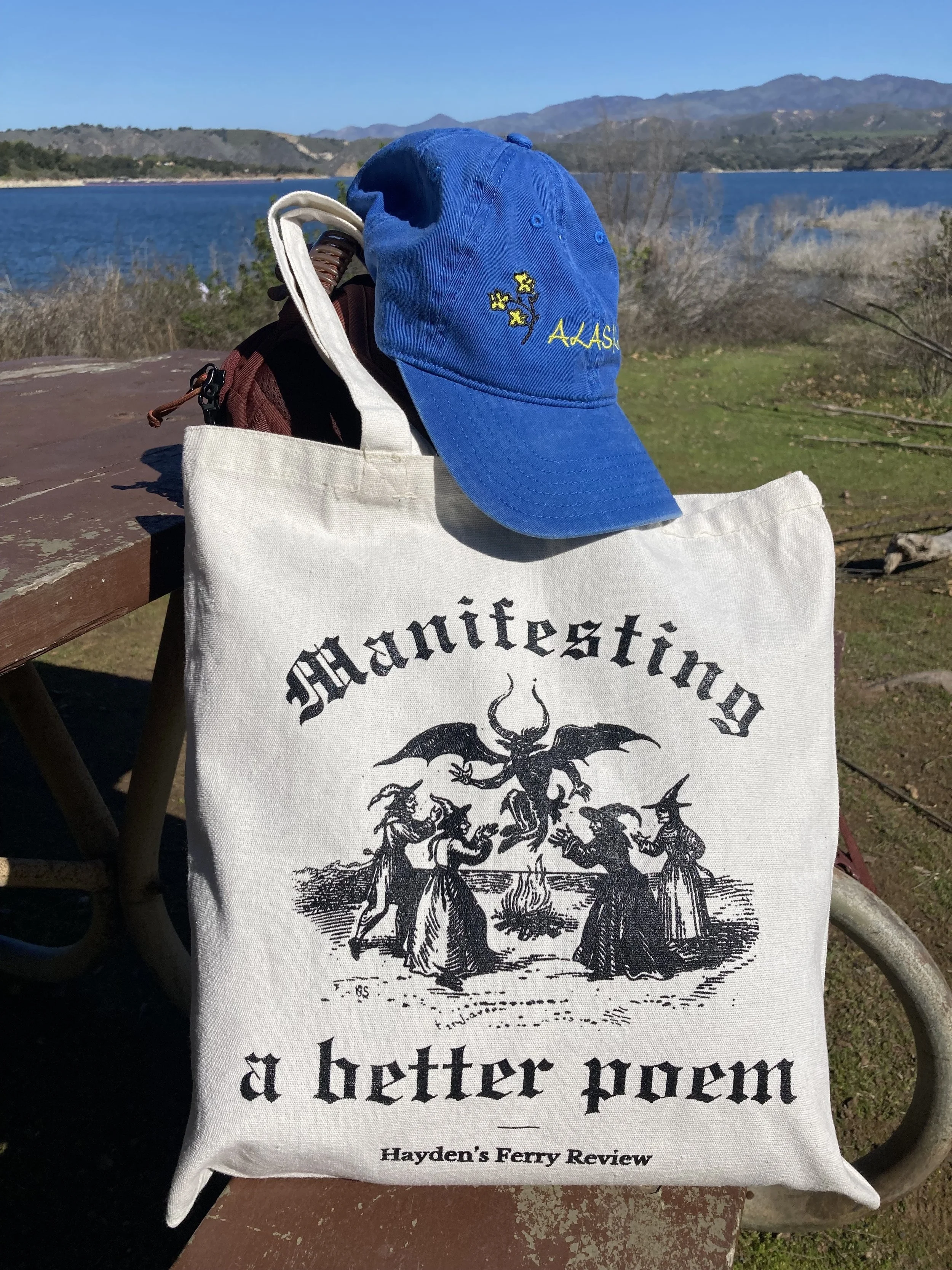A blue baseball cap rests on the Manifesting a Better Poem tote bag in front of the ocean.
