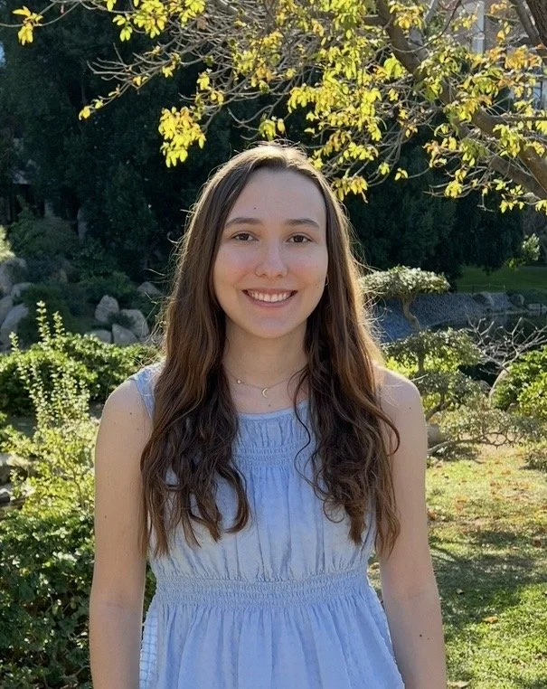 Photo of Hannah Palmisano in blue top, standing in front of ourdoor landscape, and smiling directly at camera.