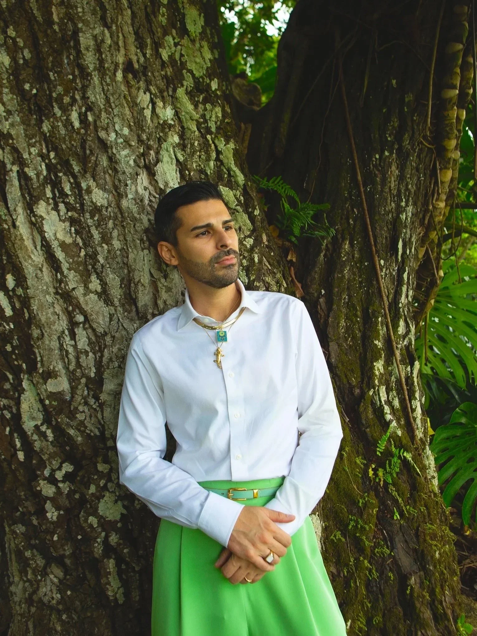 John leans against a tree and looks into distance while wearing white button up shirt and bright green pants.