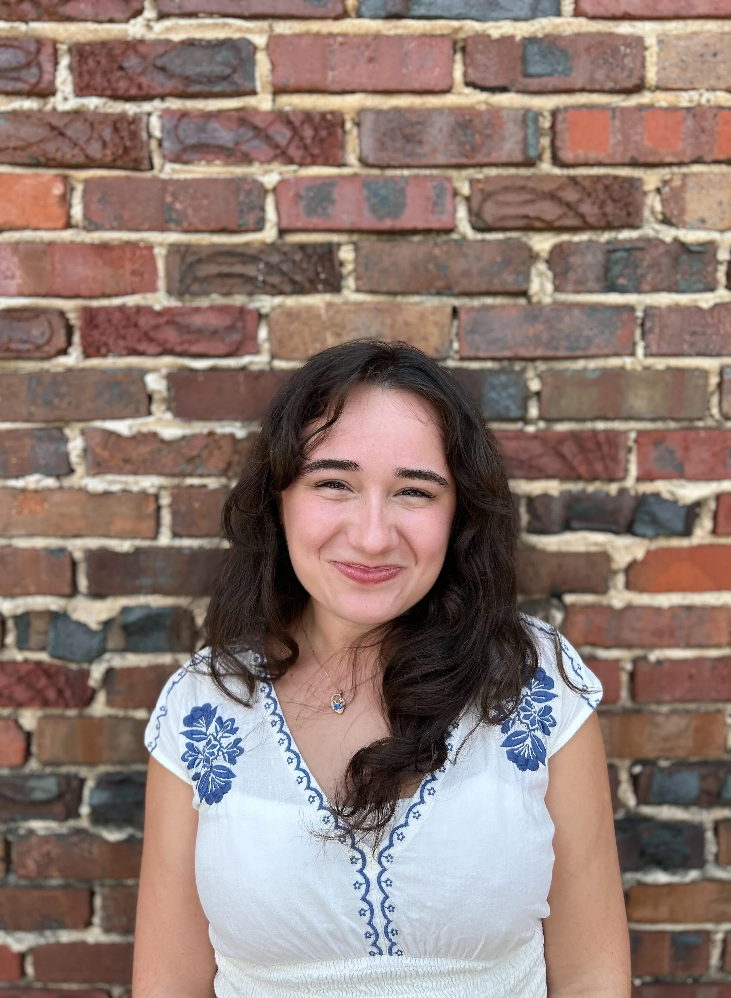 Headshot of Sarah Scarcliff in white and blue top standing in front of brick wall and smiling at camera.