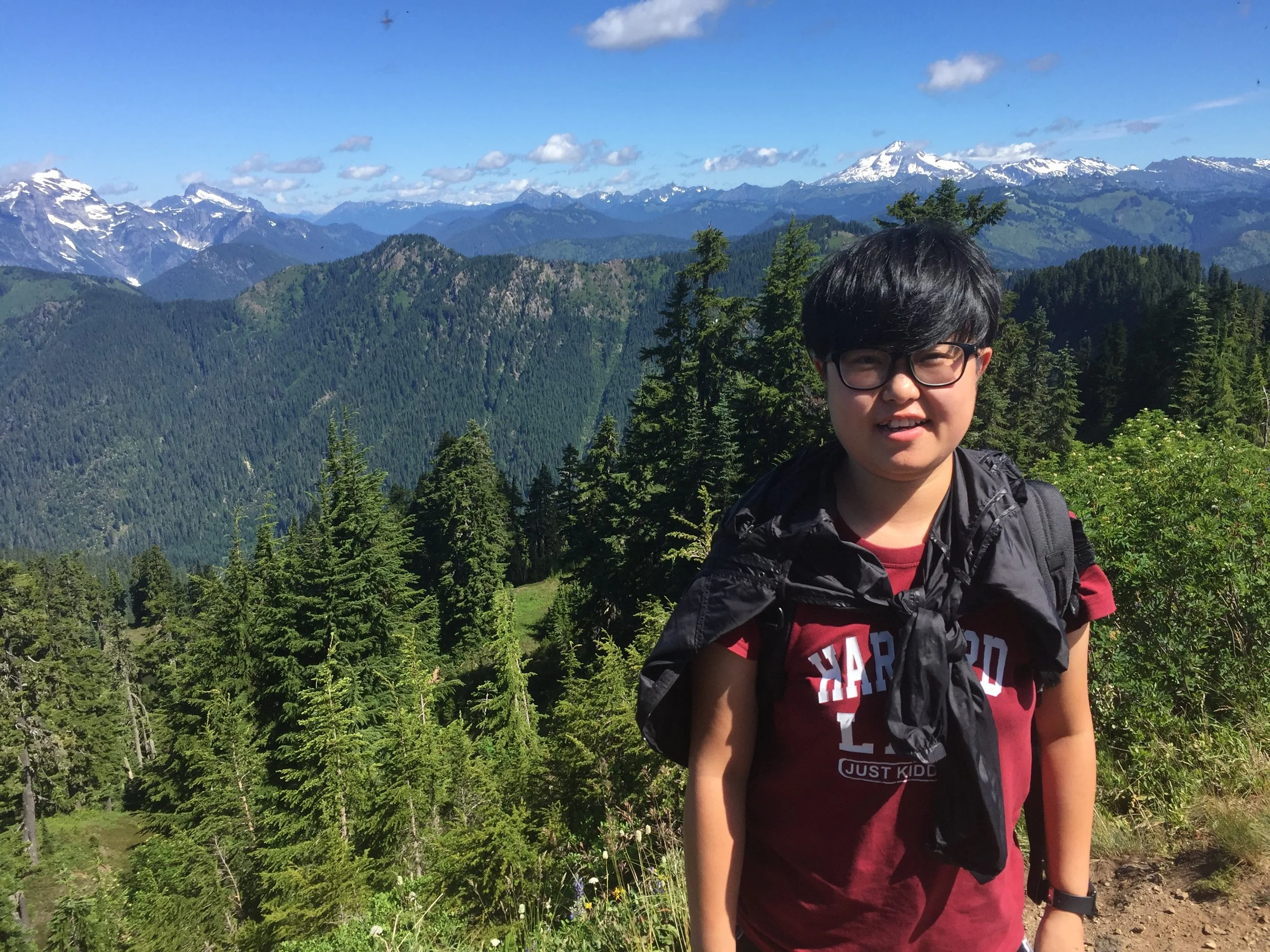 Chris poses in front of an outdoor landscape of pine trees, mountains, and clouds.