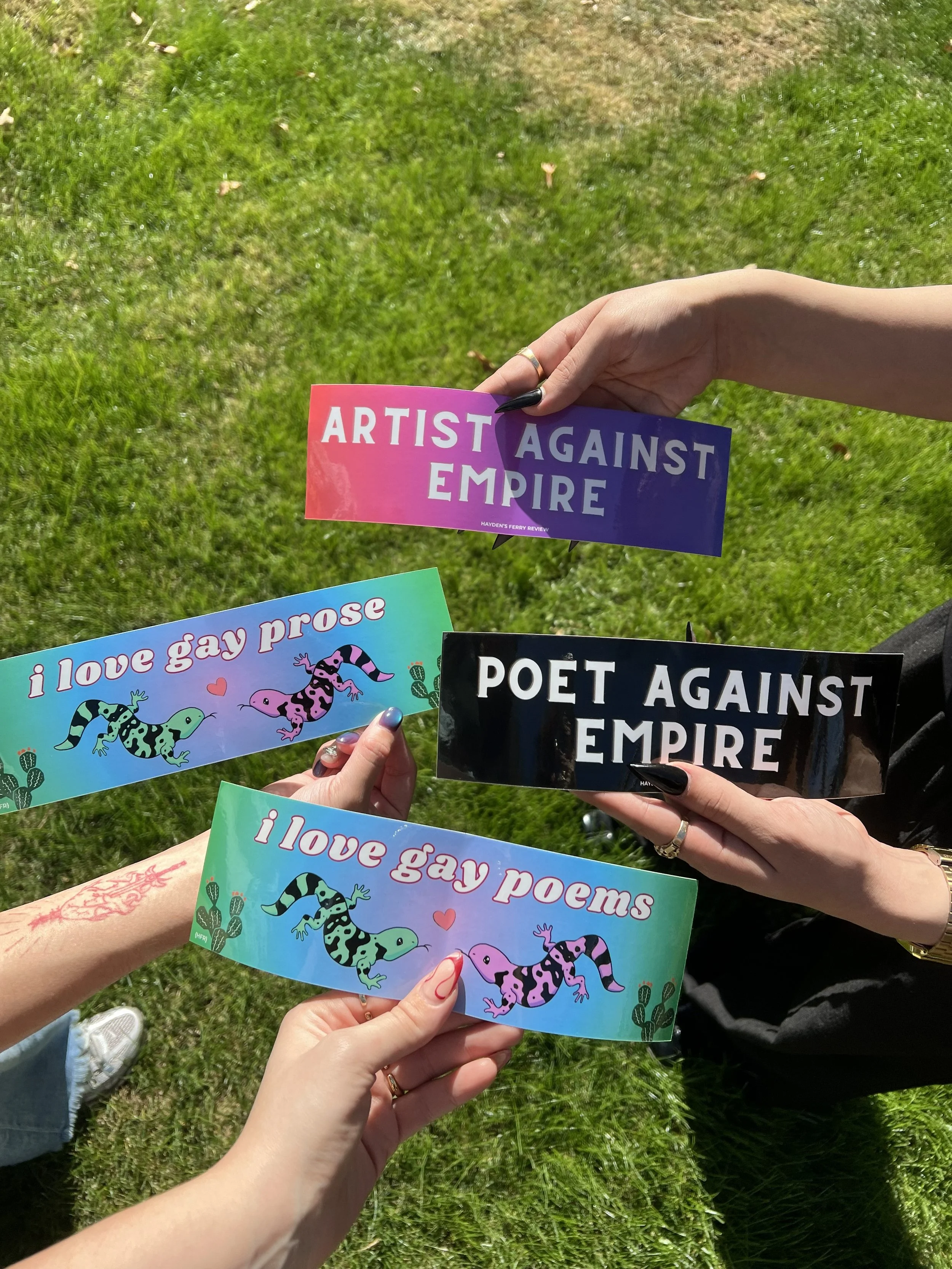 Hands hold four different bumper stickers in front of green grass.