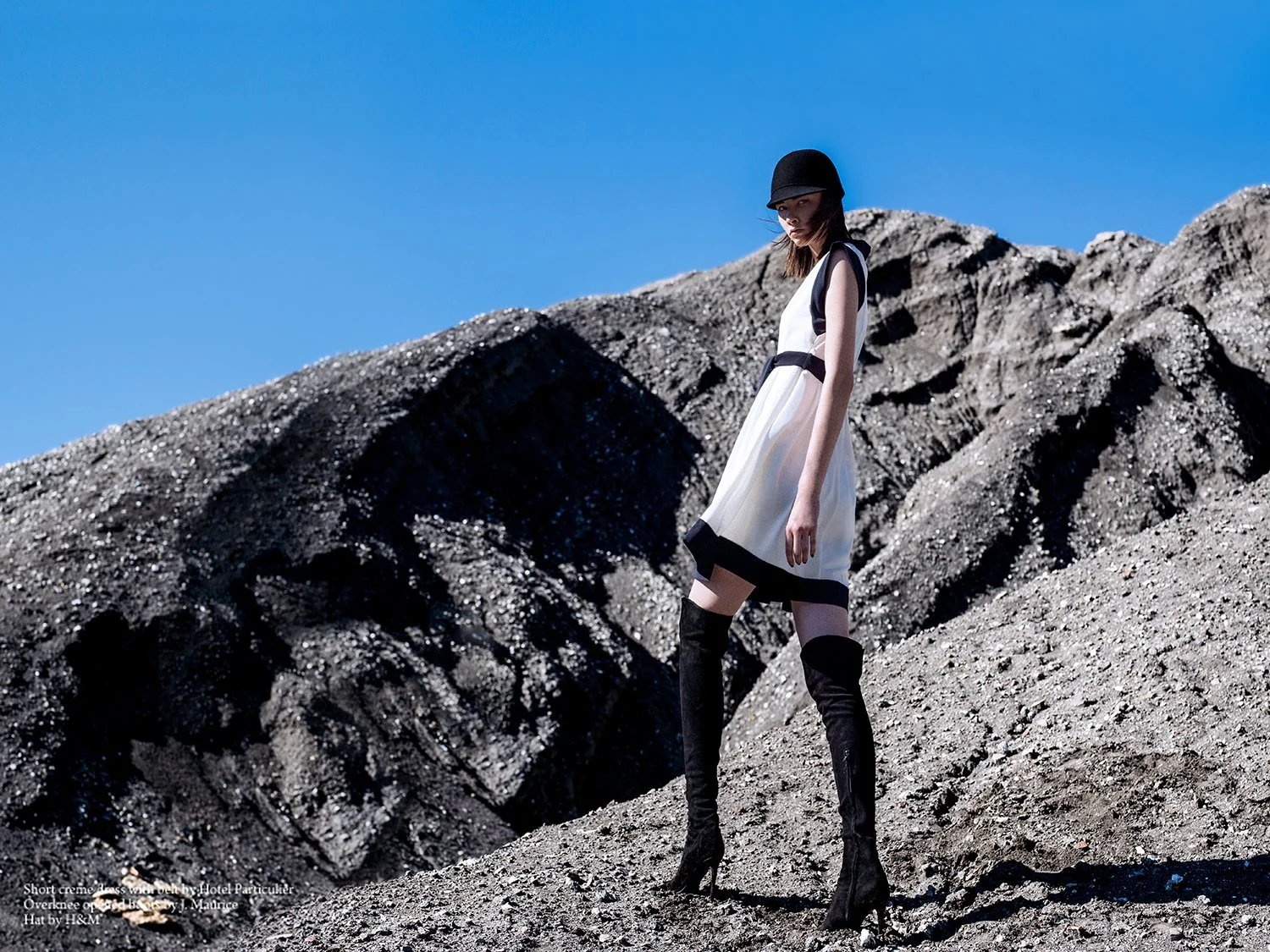 A woman standing on rocky terrain under a clear blue sky, wearing a white and black dress, a black hat, and black thigh-high boots.