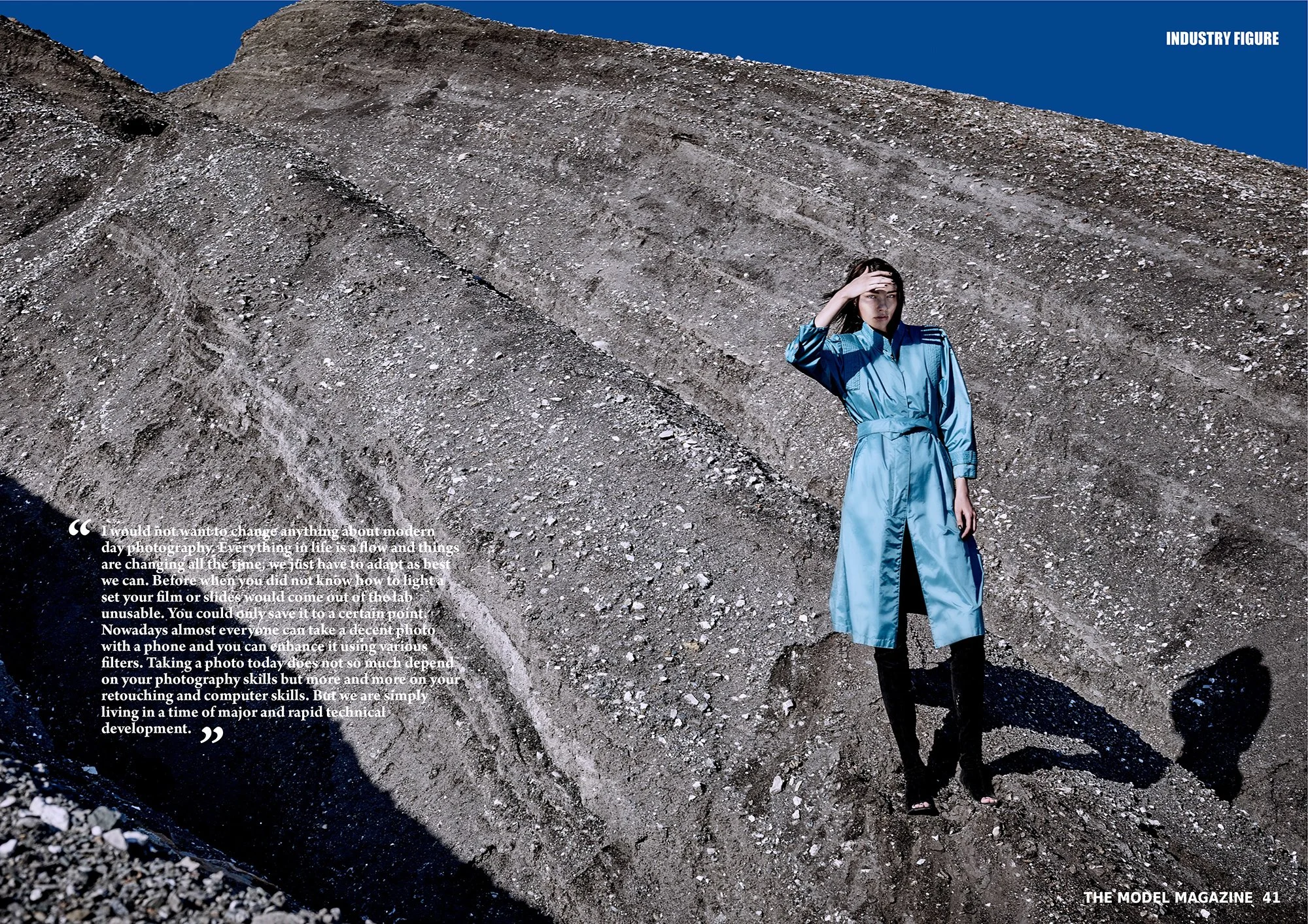 Location fashion photography by Daniella Hehmann, featuring a model in a blue trench coat. She poses with one hand over her forehead, looking into the distance. The image is on a page of The Model Magazine, featuring a quote in the lower left corner.