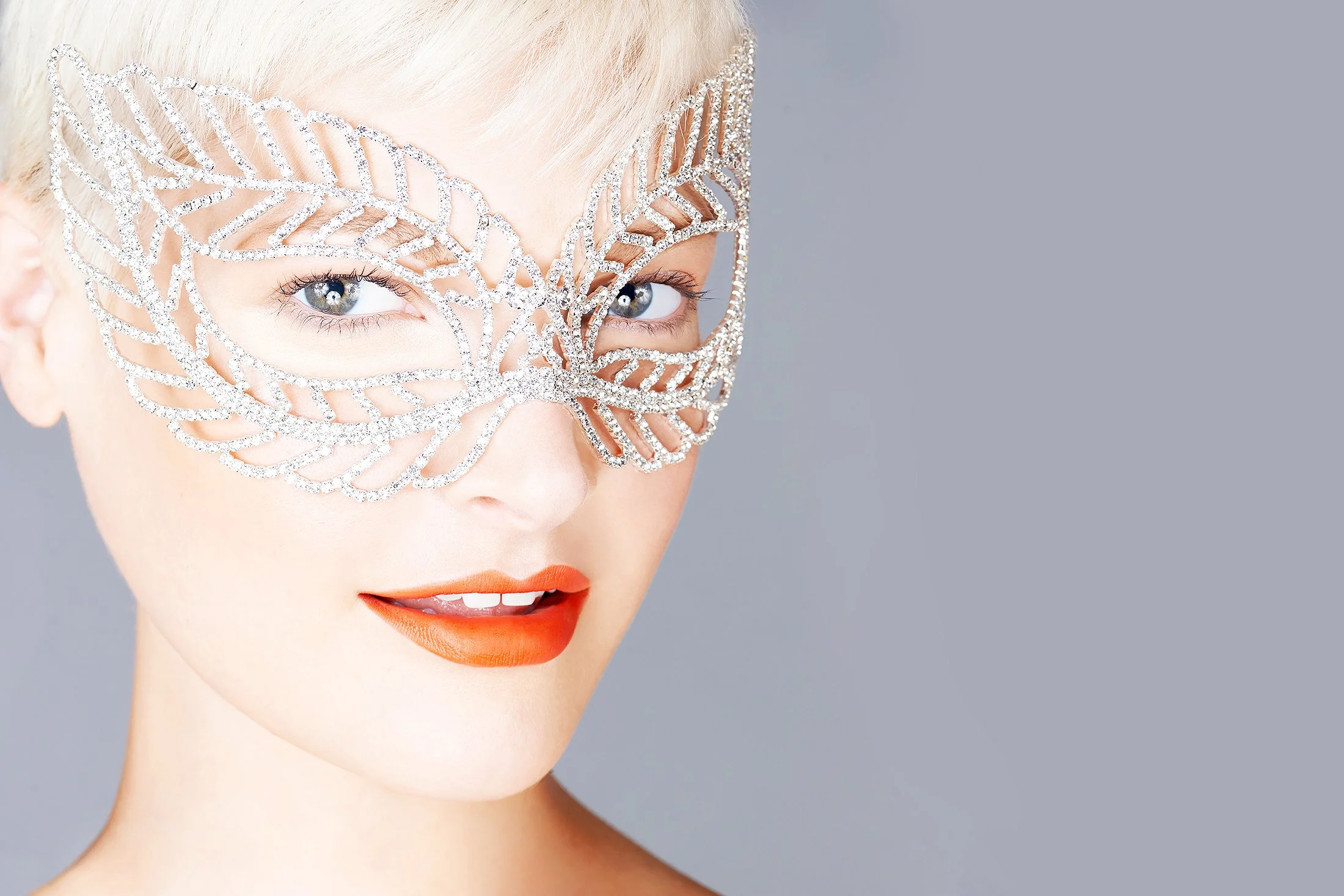 Luxury advertising photography by beauty photographer Daniella Hehmann. Close-up portrait of a blonde model wearing an intricate crystal-embellished masquerade mask and bold orange lipstick. The lighting is clean and professional, utilizing soft-box 