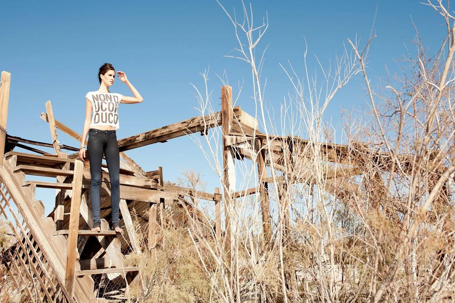 Industrial fashion editorial by photographer Daniella Hehmann for Eugene Jeans featuring a model in dark slim-fit denim standing on a weathered wooden structure amidst the arid landscape of Salton Sea.
