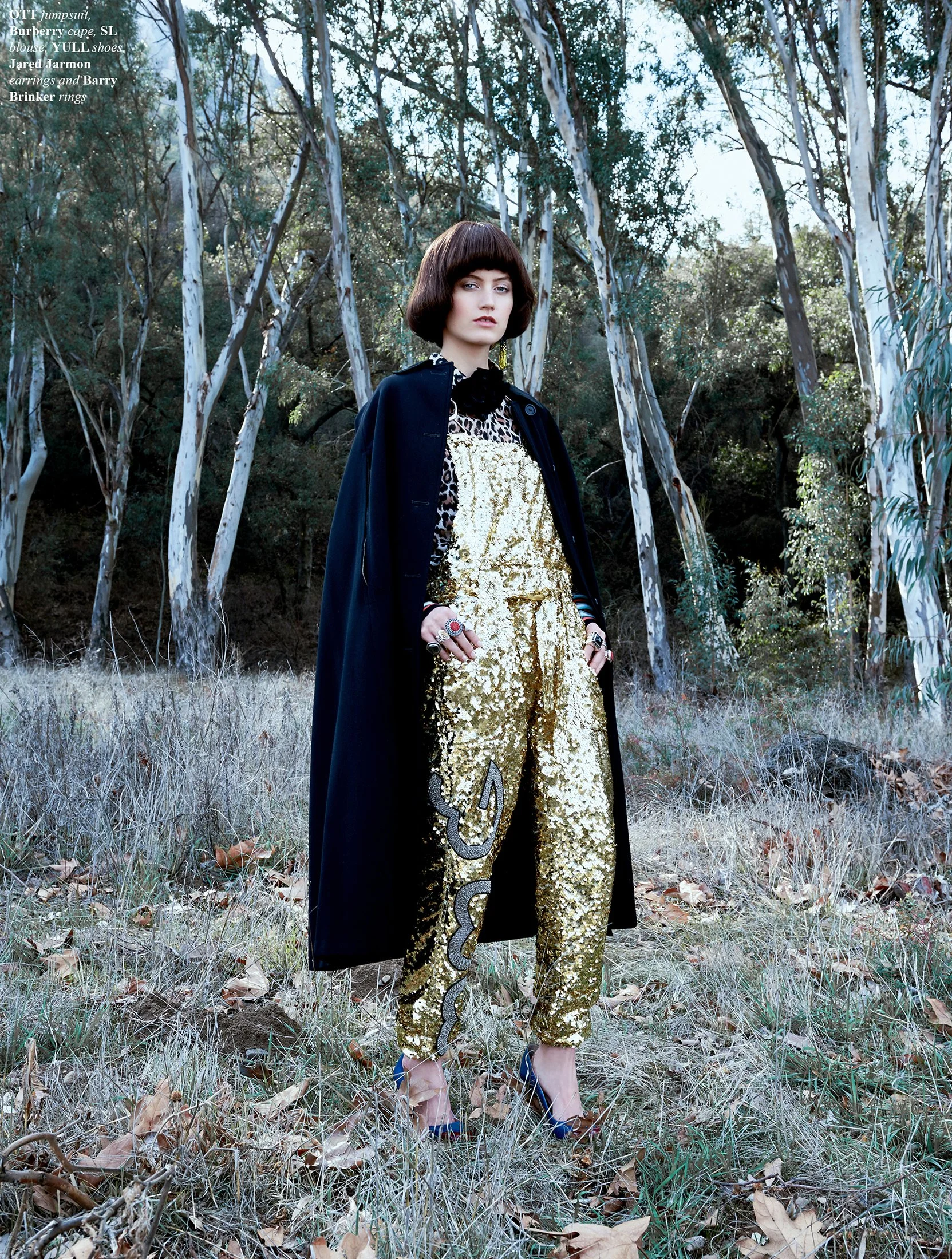 A woman standing in a forested area wearing a black coat, leopard print top with black accents, and gold sequined pants with a snake print detail, blue high heels, and multiple rings.