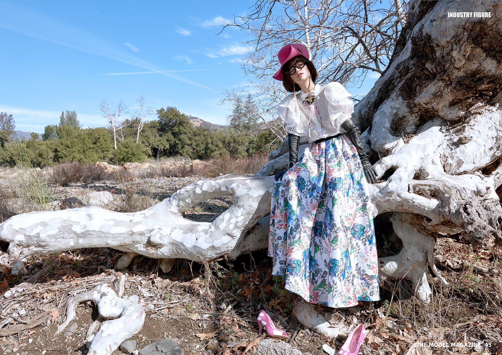 Cinematic outdoor fashion photography by Daniella Hehmann, featuring fashion model posing outdoors on a dead tree, wearing a pink hat, black glasses, a white blouse with puffed sleeves, a floral skirt, black gloves, and pink high heels on the ground.