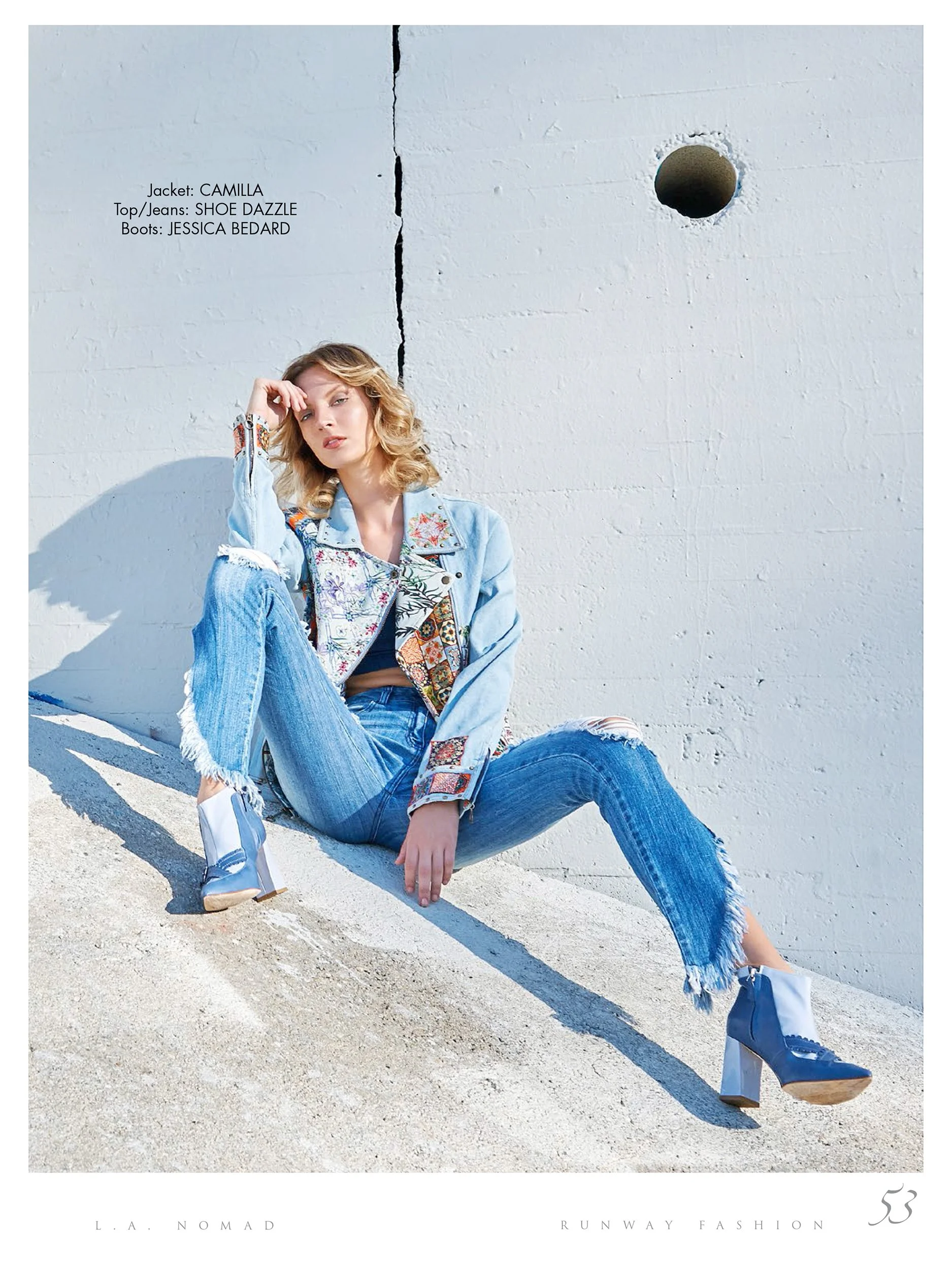 A young woman sitting on a sloped concrete surface against a white wall with a large crack. She wears a patterned denim jacket, ripped jeans, and blue and white high-heeled boots. Her blonde hair is styled in loose waves, and she is gazing towards th