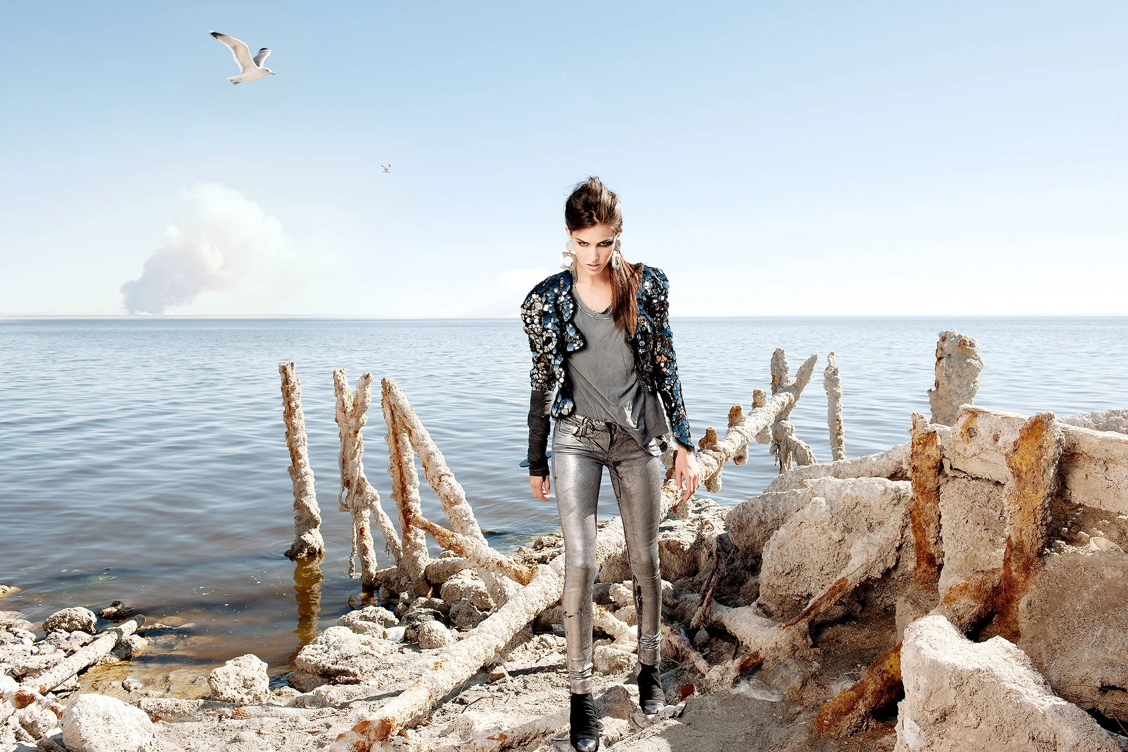 A young woman standing on a rocky shore near the ocean with salt-encrusted, broken wooden beams. Seagulls flying in the clear sky, with calm water and a cloud in the background.