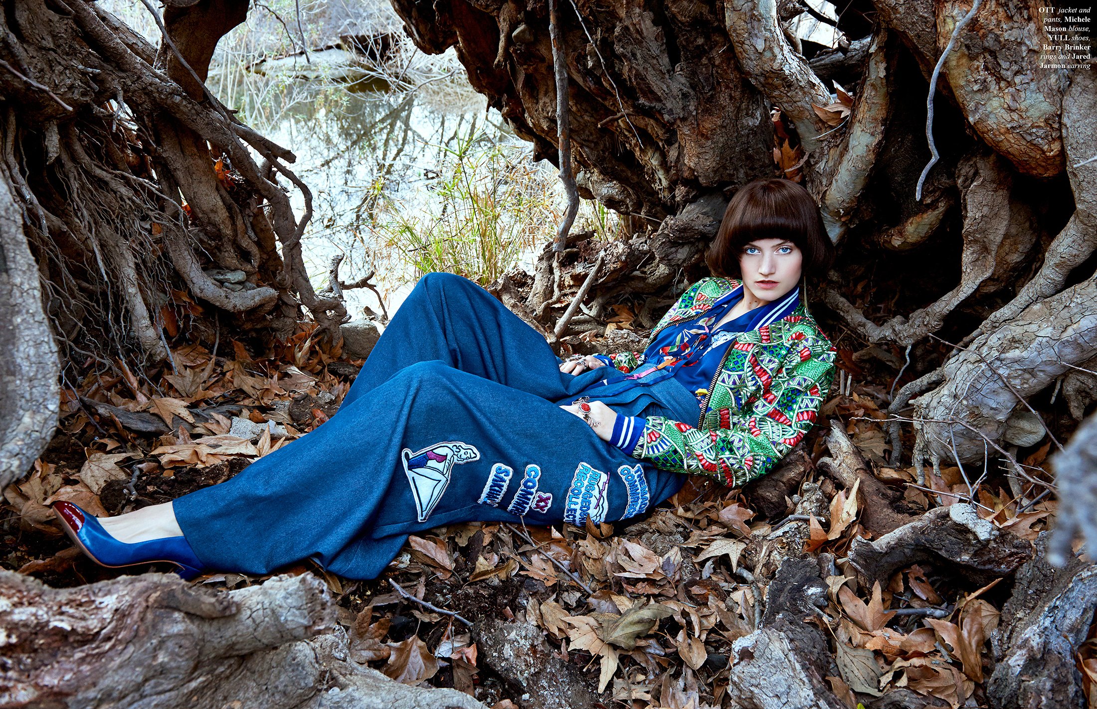 Atmospheric fashion editorial shot by photographer Daniella Hehmann in a Los Angeles forest. The model is reclining on a bed of dry leaves among tree roots, wearing a vibrant patterned jacket and wide-leg blue trousers. 