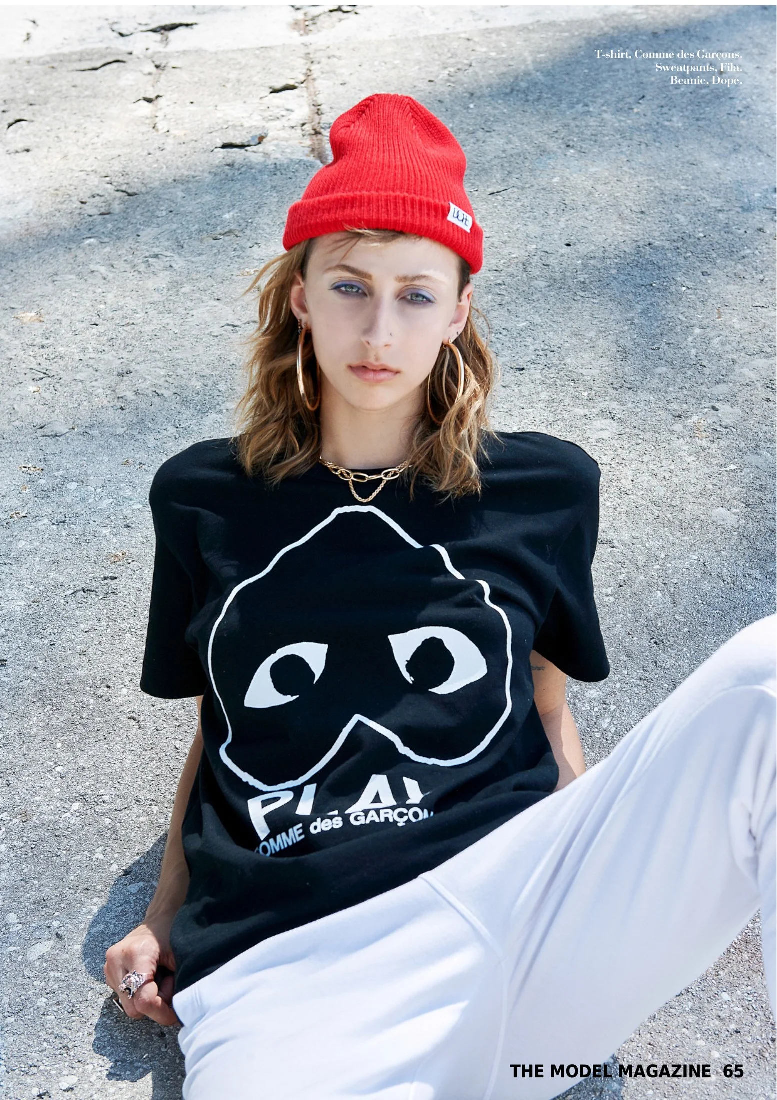 Edgy streetwear photography by photographer Daniella Hehmann shot on location in L.A., featuring a model in a black heart-logo t-shirt and red beanie, reclining on sun-bleached concrete.