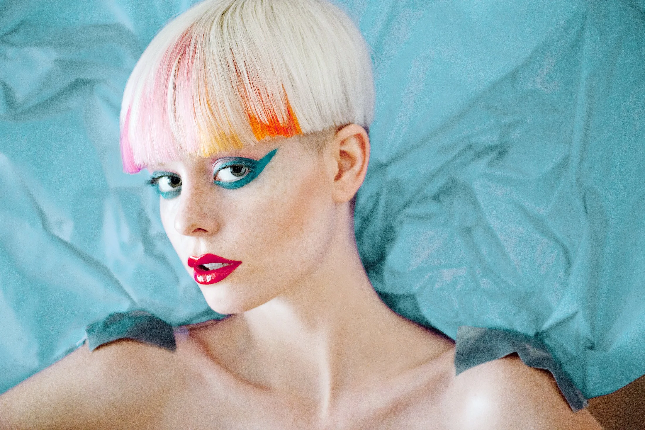A woman with short platinum blonde hair with pink and orange tips, wearing vibrant makeup including blue eyeshadow, bold blue eyeliner, and bright red lipstick, lying on crumpled light blue paper.