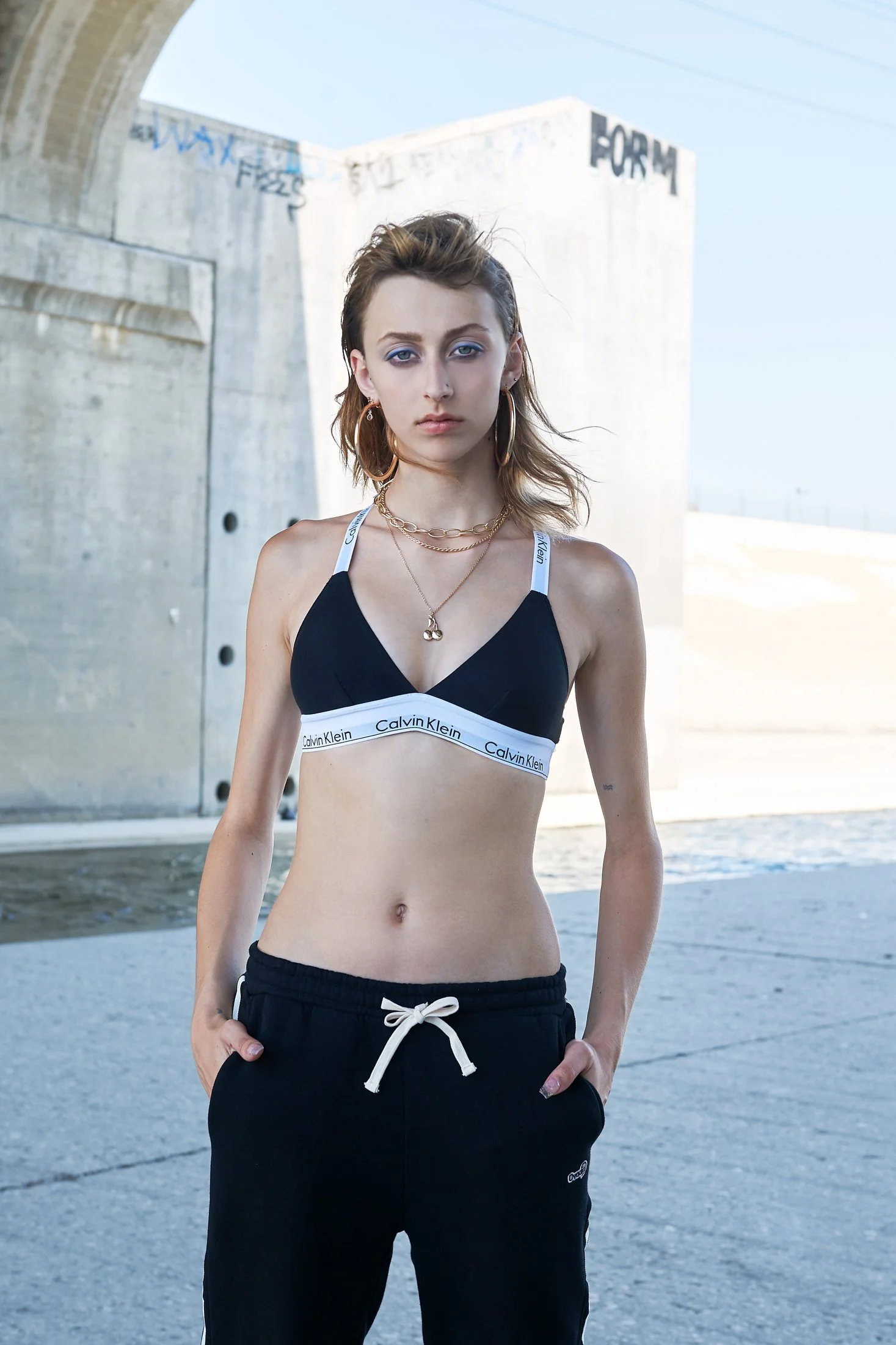 A young woman with light skin and shoulder-length brown hair stands outdoors near a concrete bridge. She wears a black Calvin Klein bralette, black sweatpants with a white drawstring, and gold jewelry, including hoop earrings and layered necklaces. S