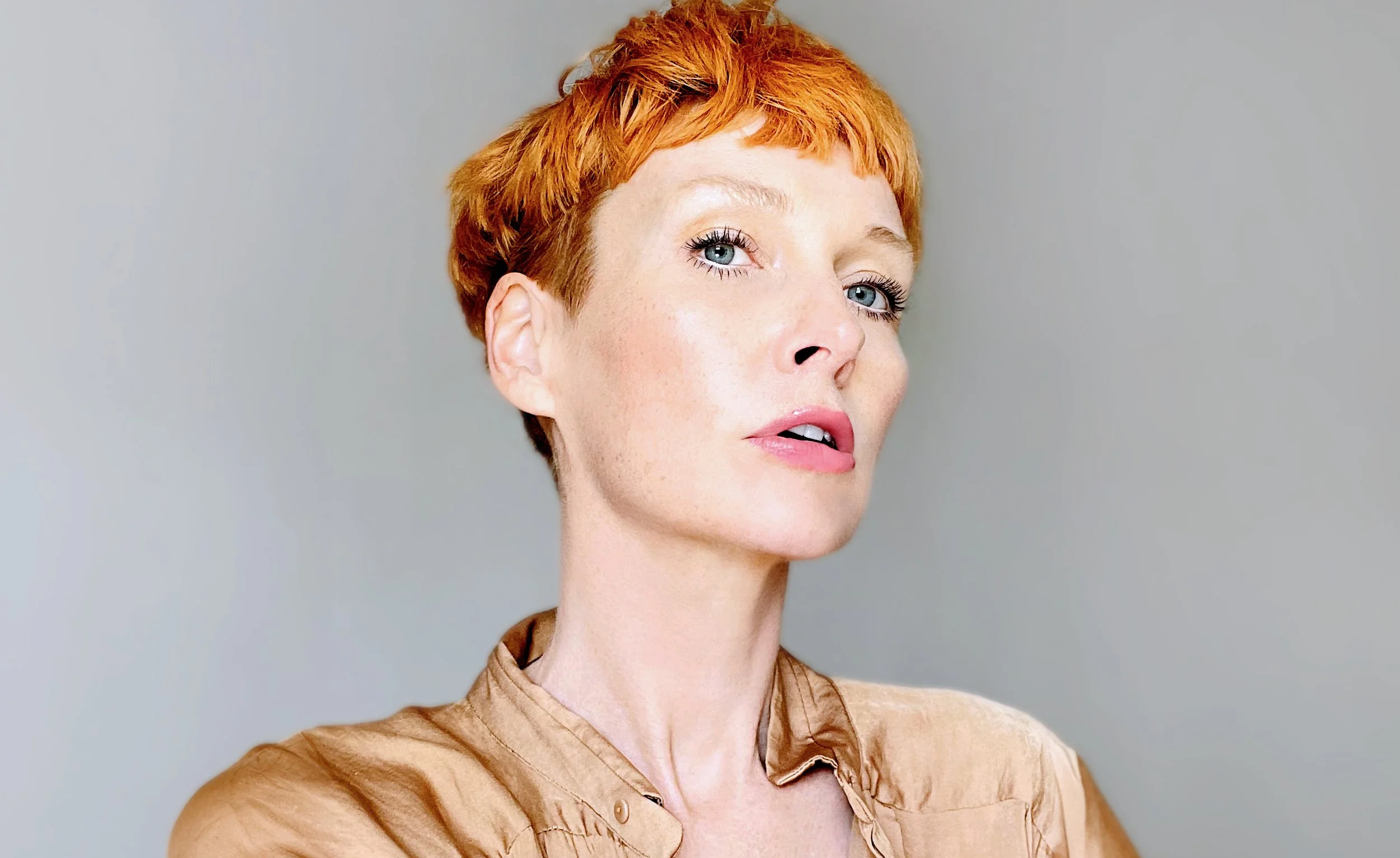 A woman with short red hair and blue eyes, wearing a tan top, posing against a plain light-colored background.