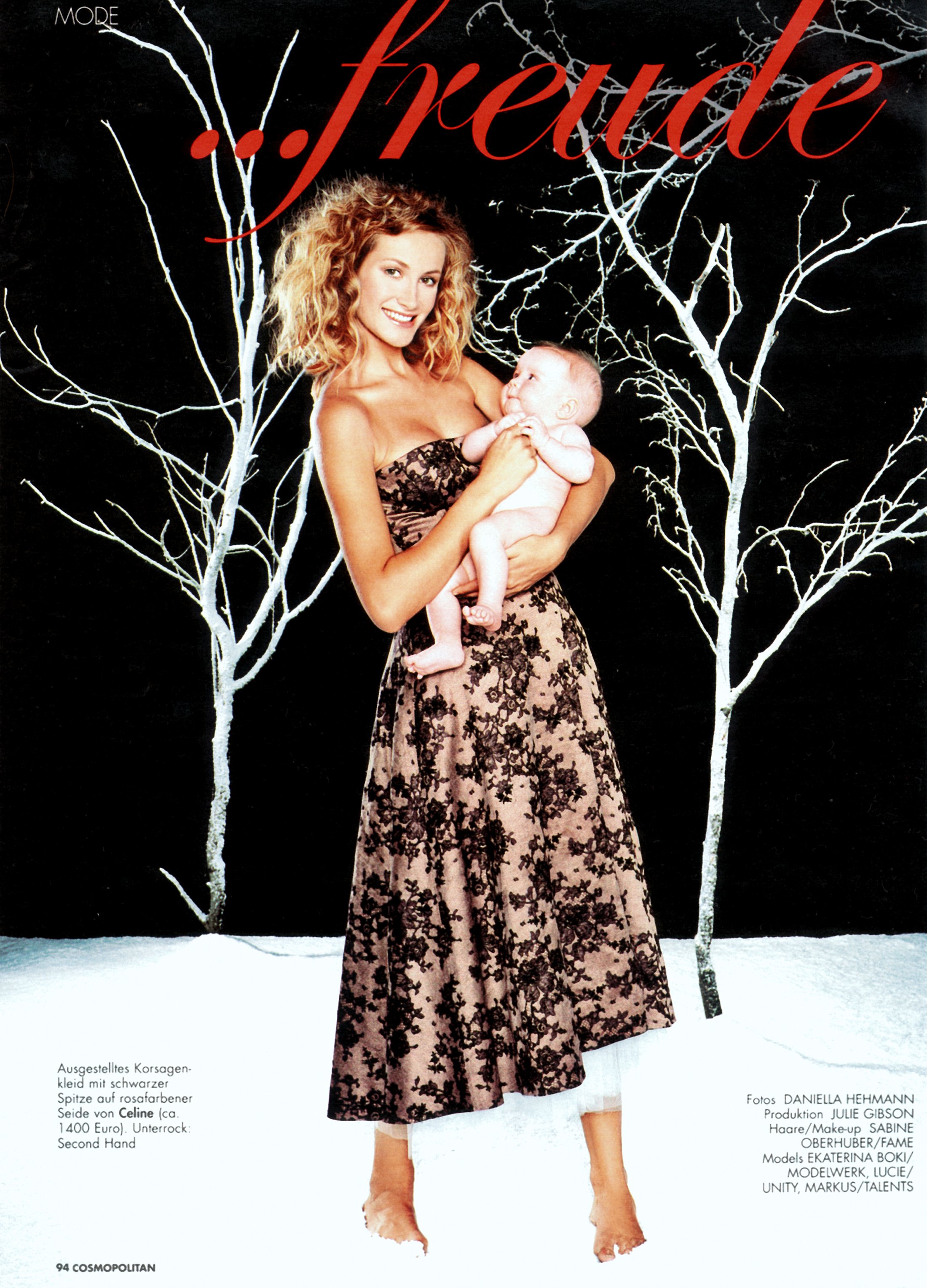 A poignant fashion editorial shot for German Cosmopolitan by Daniella Hehmann Photography featuring a model in a floral lace dress holding an infant against a black background with minimalist white tree art.