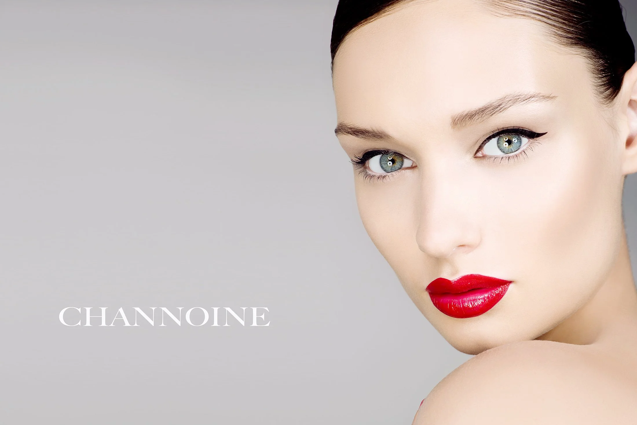 Close-up of a woman with bright red lipstick, blue eyes, and dark hair, looking over her shoulder at the camera. The background is light gray, and the word 'Channoine' appears in white text on the left side.