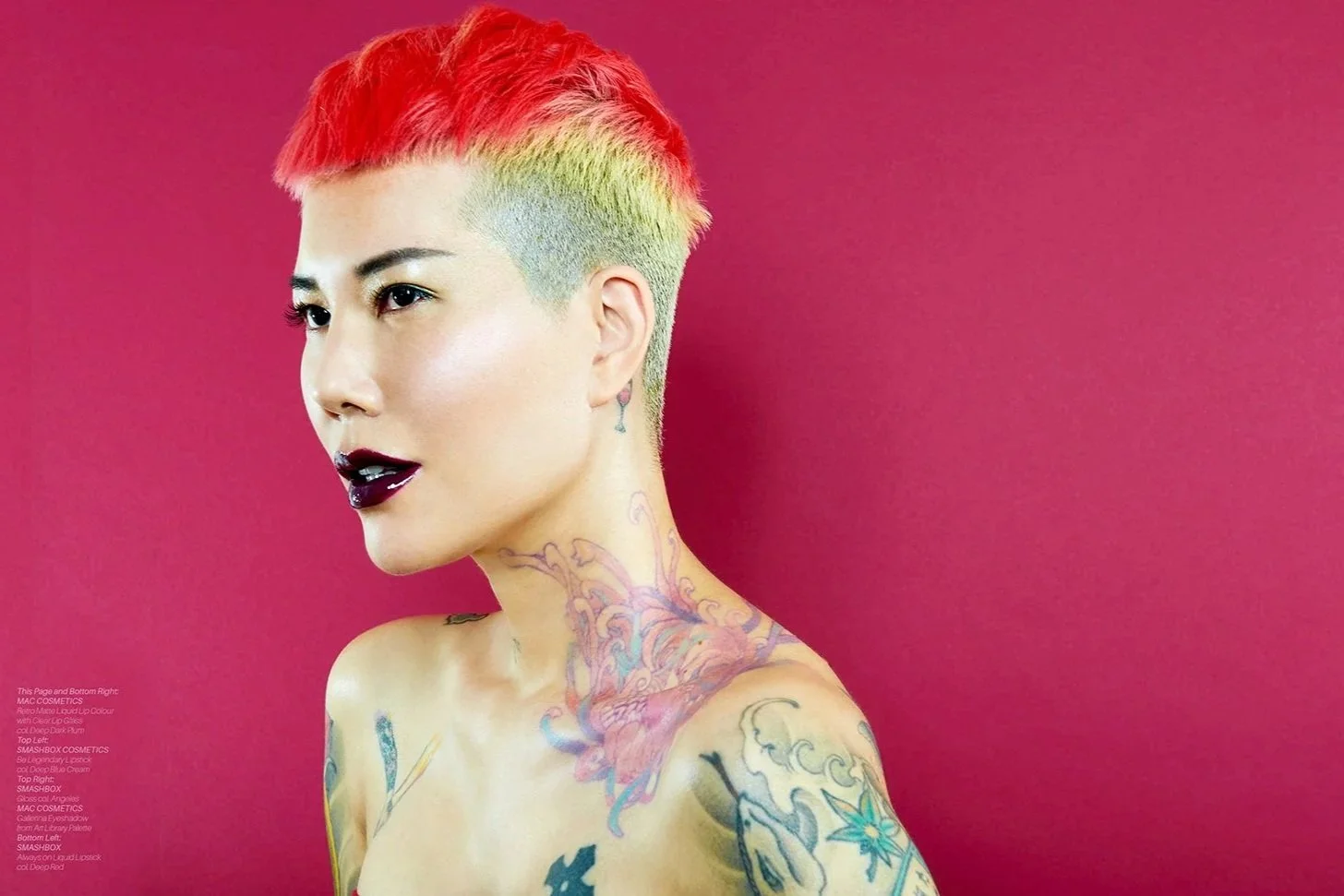 A woman with short, brightly colored hair, tattoos on her neck and shoulder, dark lipstick, and a light complexion.