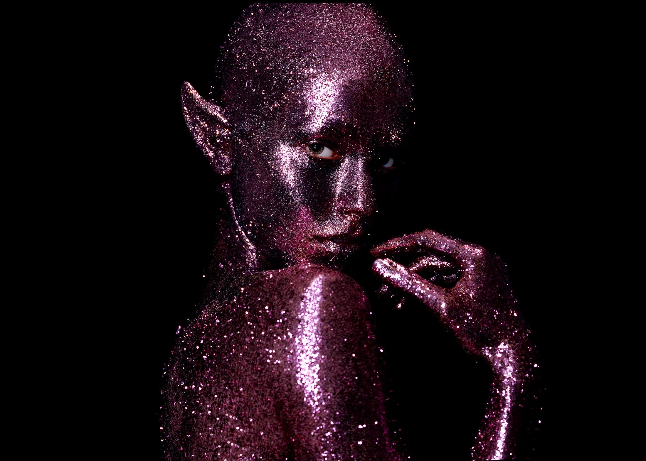 A person with glittery pink and purple body paint, glitter makeup, and striking eyes, posing against a black background.