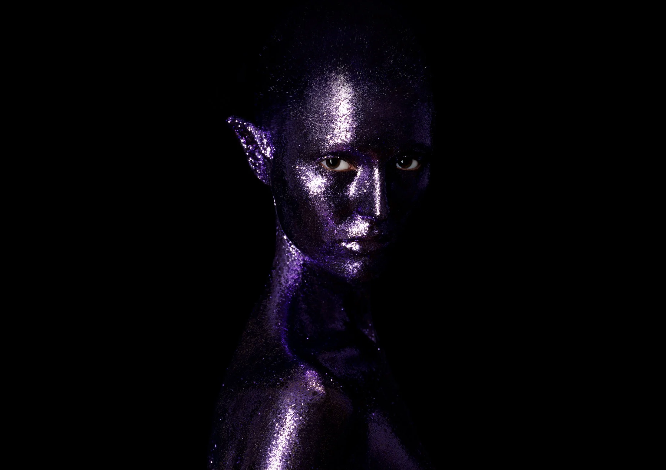 A person with purple glittered skin and elf-like ears poses against a black background.