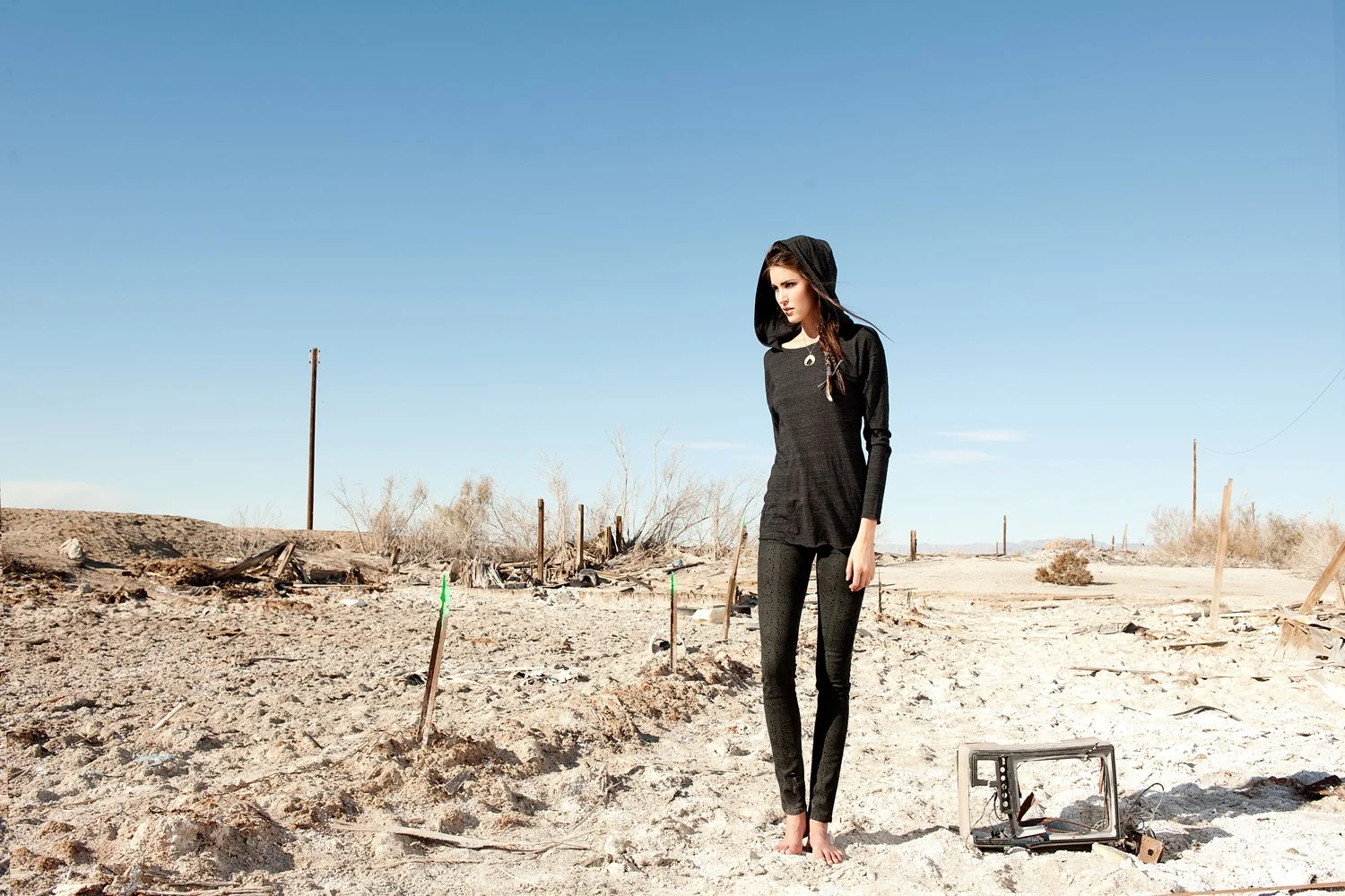 Edgy outdoor fashion photography by Daniella Hehmann Photography for Eugene Jeans showcasing a model in dark denim and a hooded top standing in the rugged sun-drenched desert landscape of the Salton Sea.