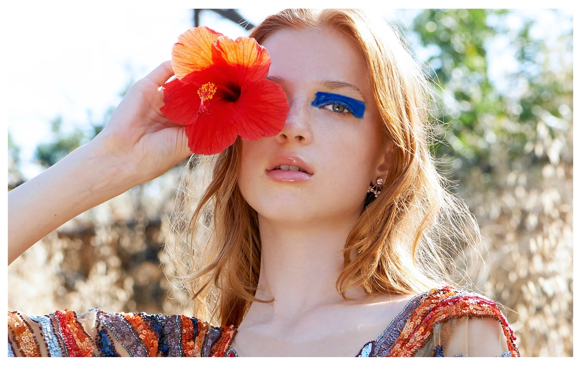 A young woman with red hair and blue eye makeup holds a red hibiscus flower near her face outdoors with blurred trees in the background.