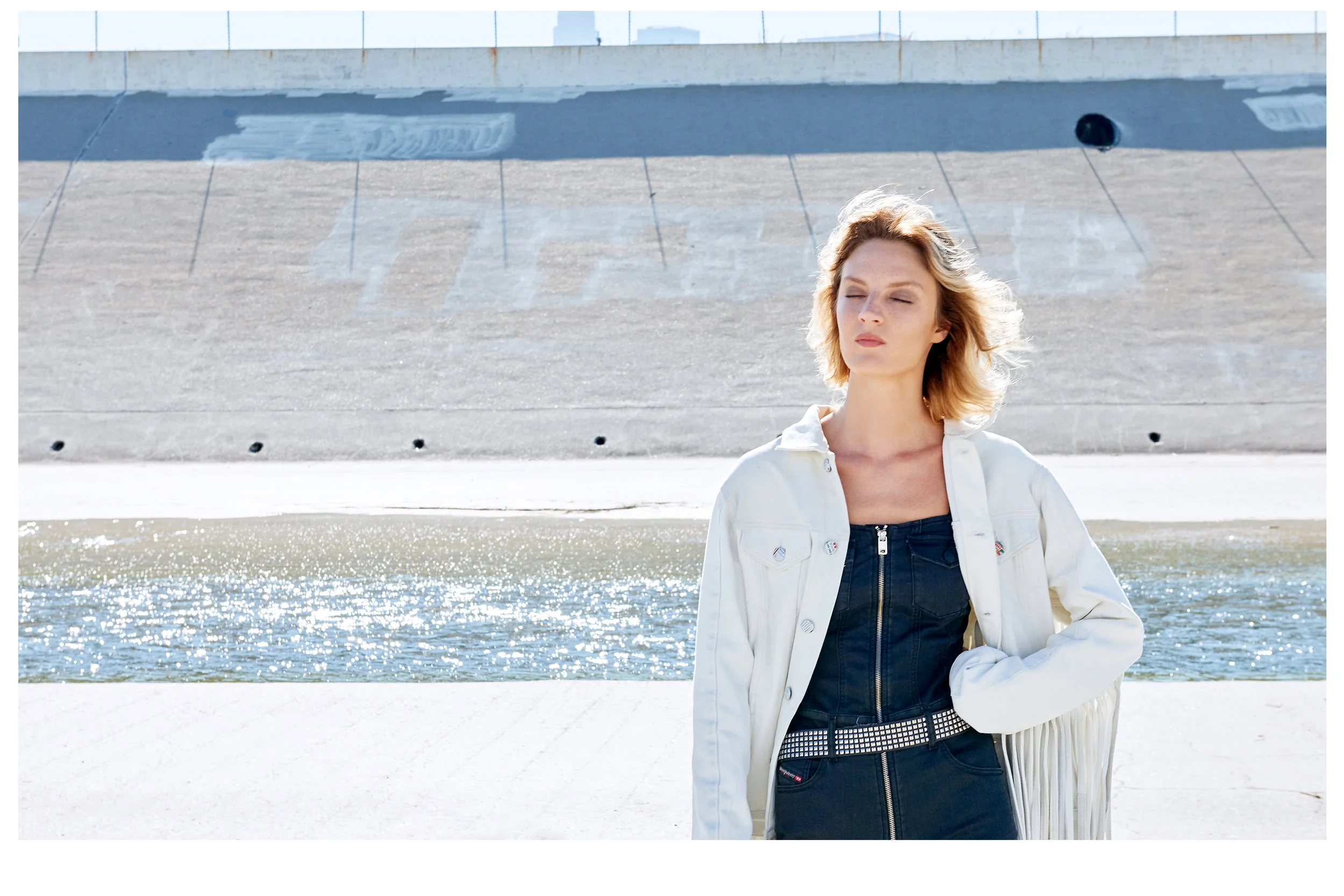 Clean fashion shot by photographer Daniella Hehmann in the Los Angeles River, featuring a model in a white denim jacket and dark jumpsuit against the minimalist concrete architecture of the riverbed.