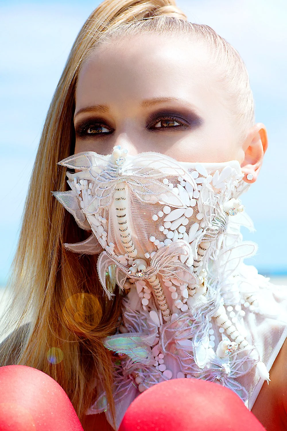 Detailed close-up fashion shot by photographer Daniella Hehmann of a model wearing an ornate, white lace and pearl-encrusted face mask with iridescent accents, paired with bold smoky eye makeup under bright, sunny light.