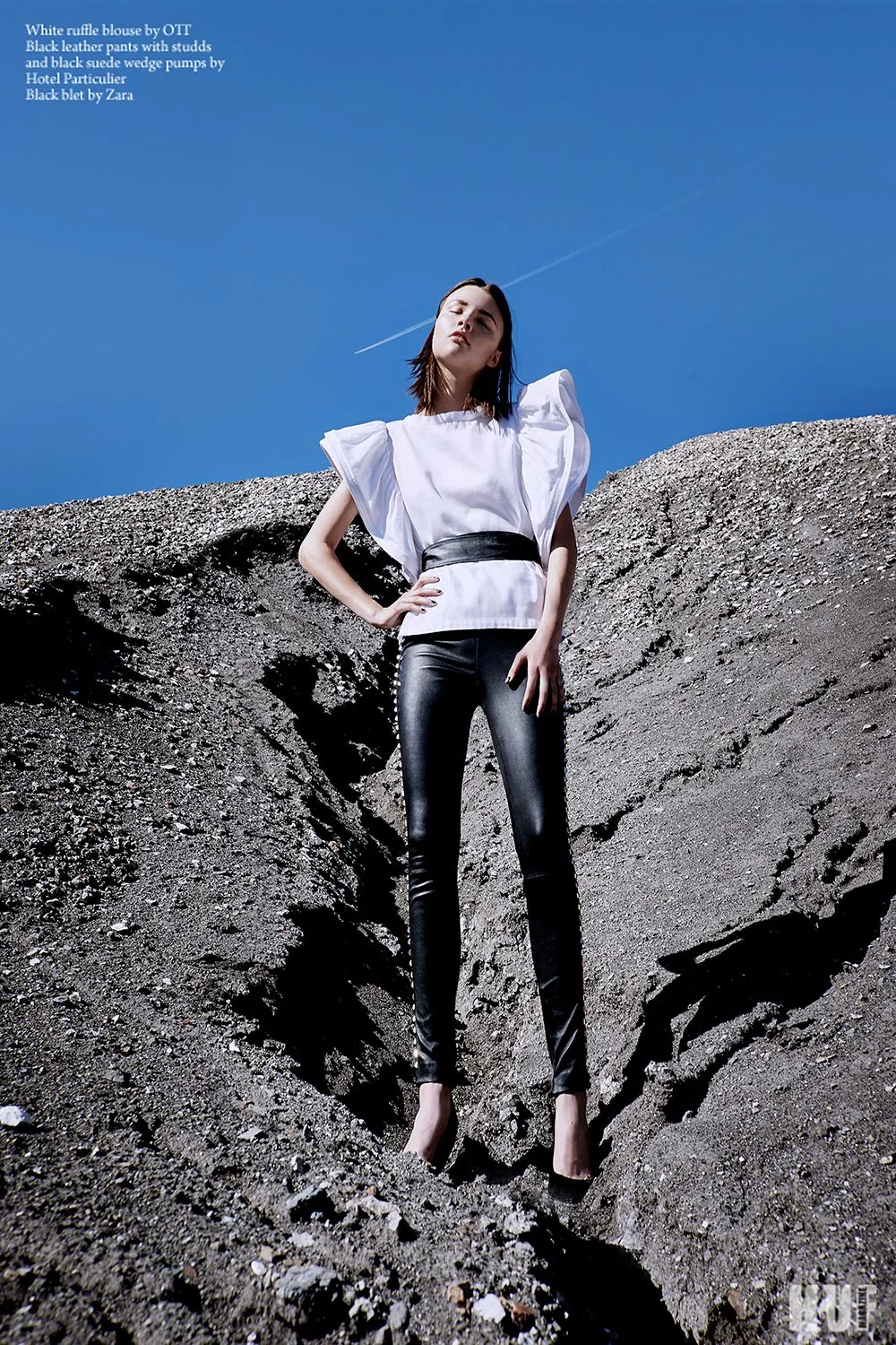 A woman wearing a white ruffle blouse by OTT, black leather pants with studs, and black wedge pumps by Hotel Particulier, stands confidently on a rocky, gray terrain under a bright blue sky with a contrail visible in the background.