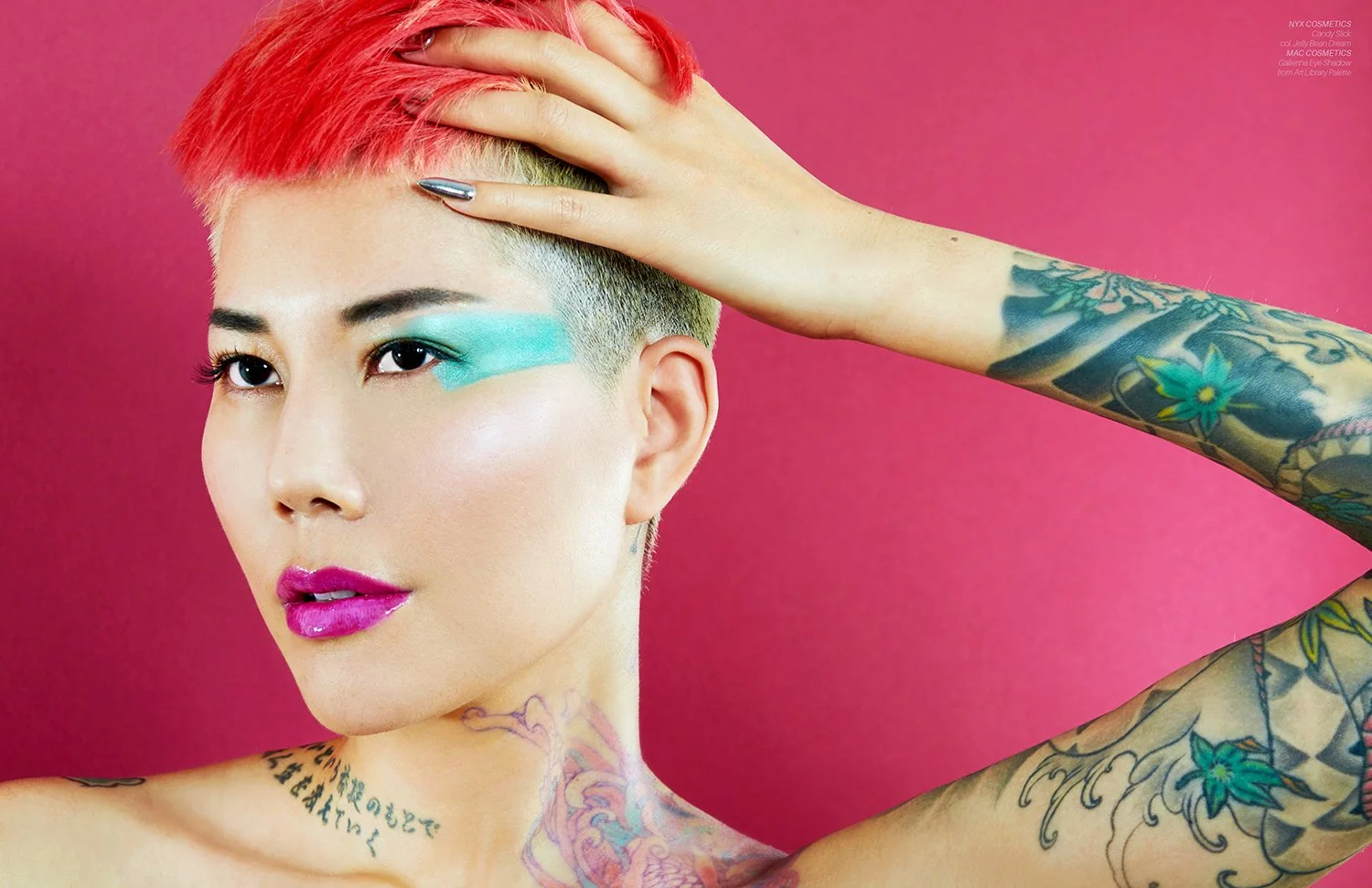 Close-up portrait of a woman with short, brightly colored hair, green eye makeup, and pink lipstick. She has tattoos on her neck and arms, and poses with her hand on her head against a pink background.