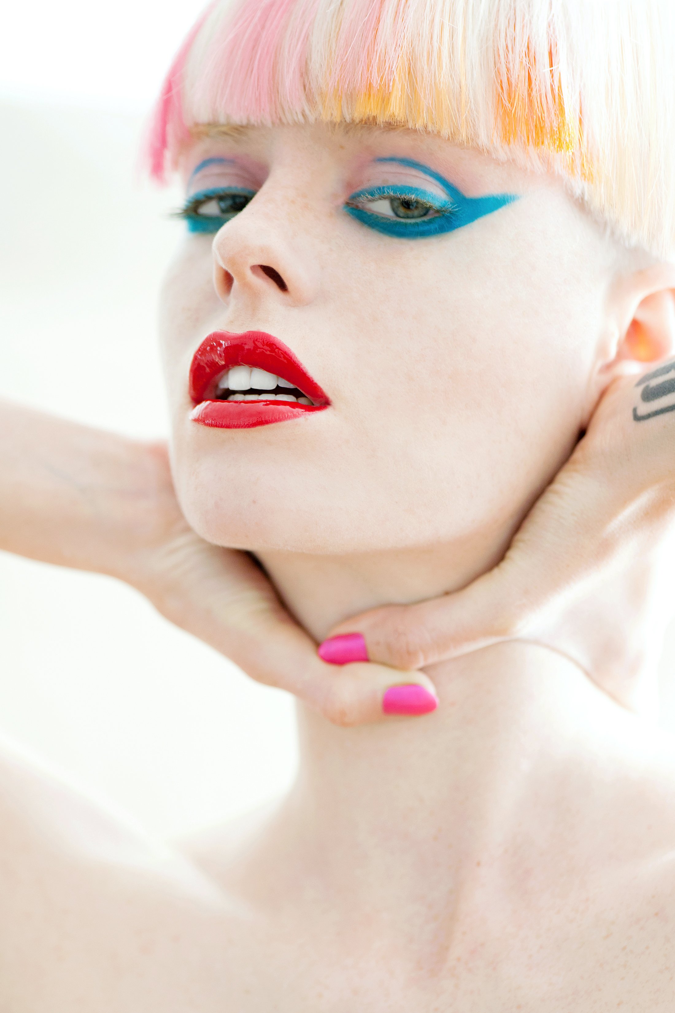 Beauty photography by Daniella Hehmann: Close-up of a woman with bold makeup, pink and yellow hair, and pink lipstick, holding her neck with one hand.