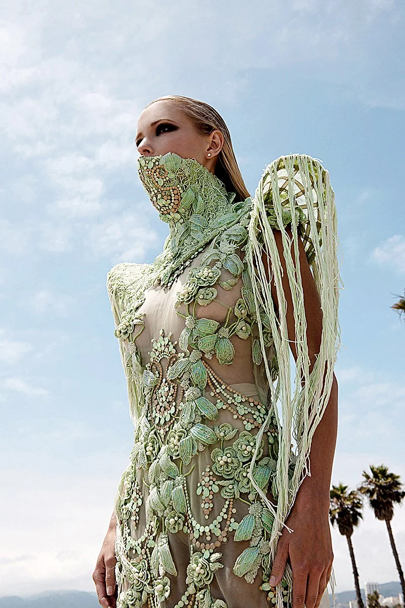 Ethereal fashion shot by photographer Daniella Hehmann featuring a model in a pale green sequined gown with a high structural neck against a clear blue sky.