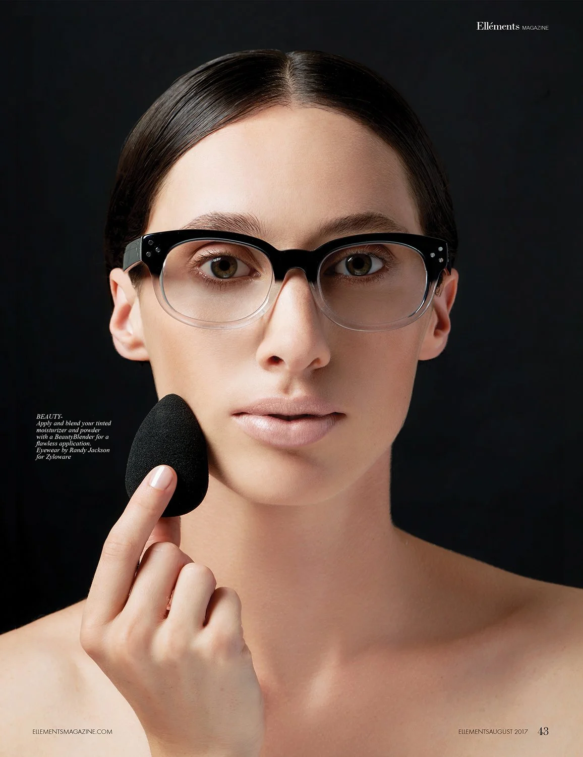 A woman with glasses and dark hair holds a rounded makeup sponge near her face against a black background.