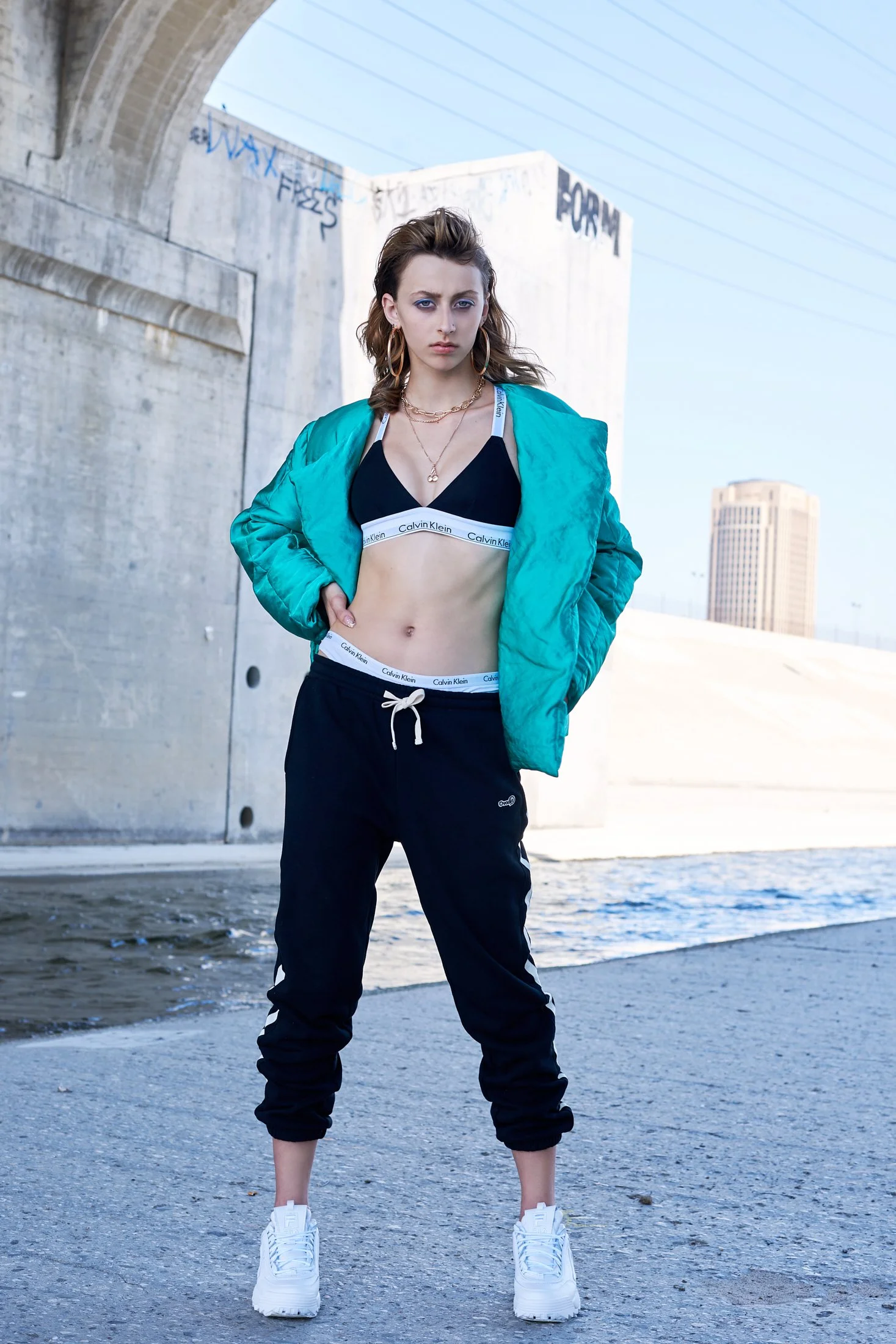 Young woman standing outdoors near a concrete bridge, wearing a black Calvin Klein sports bra and sweatpants, with a teal jacket draped over her shoulders, white sneakers, and jewelry, against urban background.