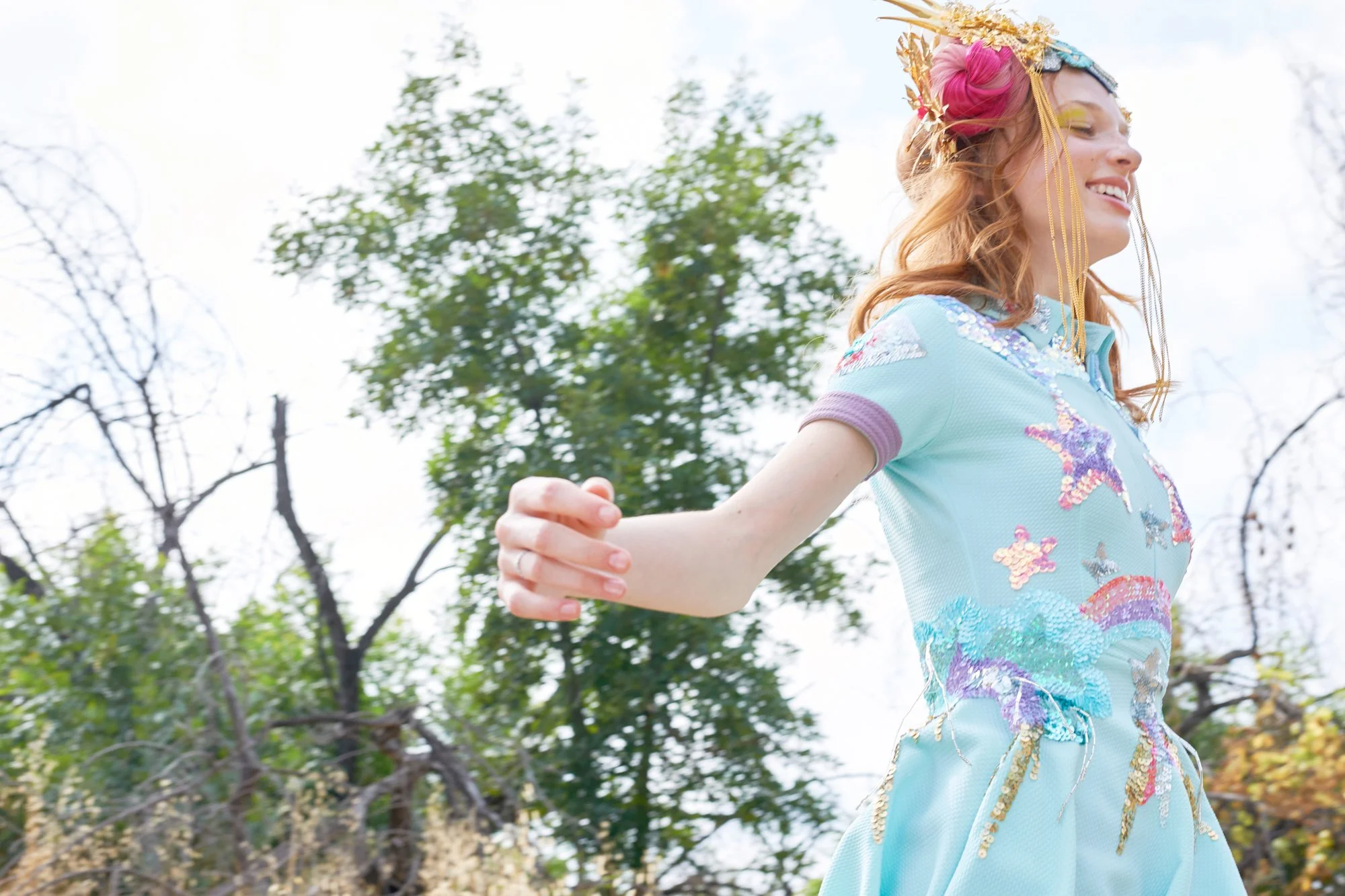 Woman with a colorful, embroidered dress and an ornate headdress, smiling and stretching her arms outdoors near trees with autumn leaves.