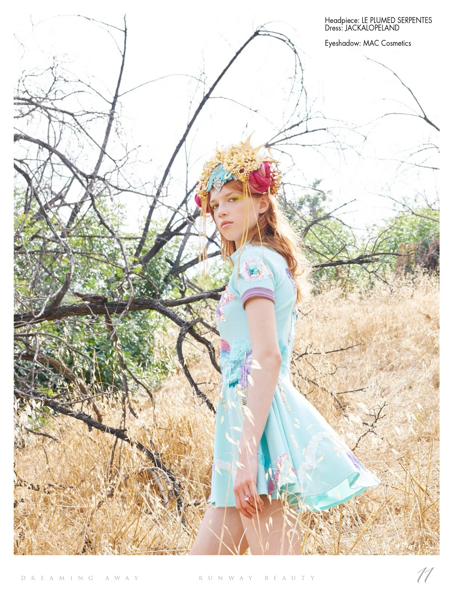 A model with light skin wears a pastel blue dress with colorful embroidery and a large, elaborate headpiece made of gold, pink, and blue accents. She stands outdoors in a dry, grassy field with leafless trees in the background, looking at photograph.
