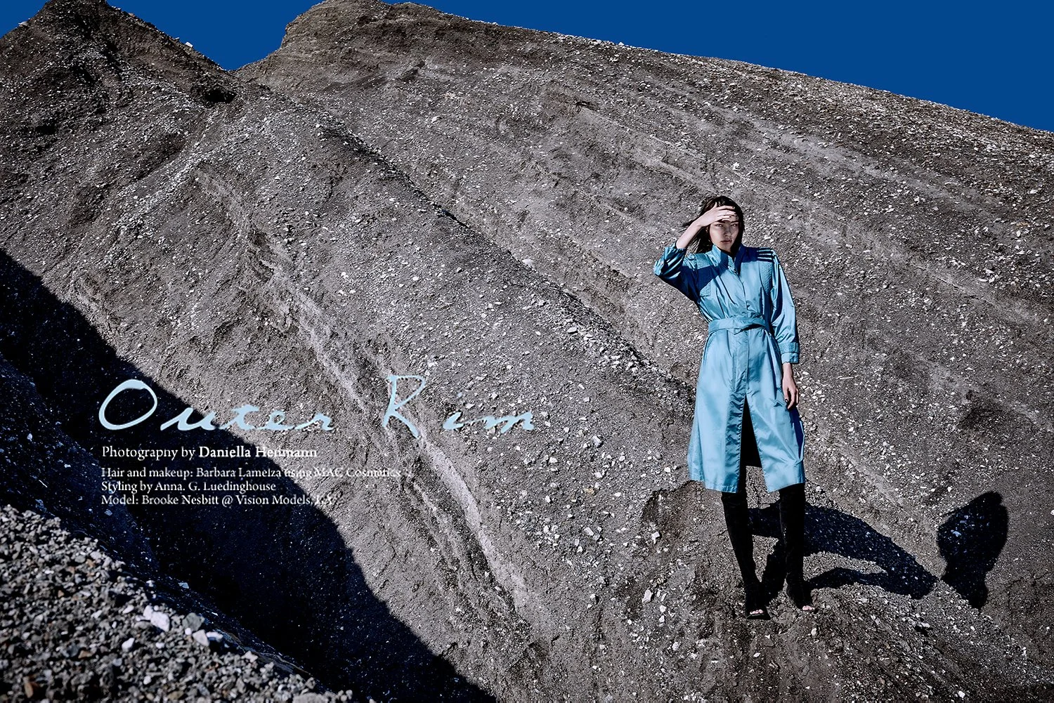 A woman in a shiny blue trench coat and black boots standing on rocky terrain, with a large dirt mound behind her and clear blue sky above.
