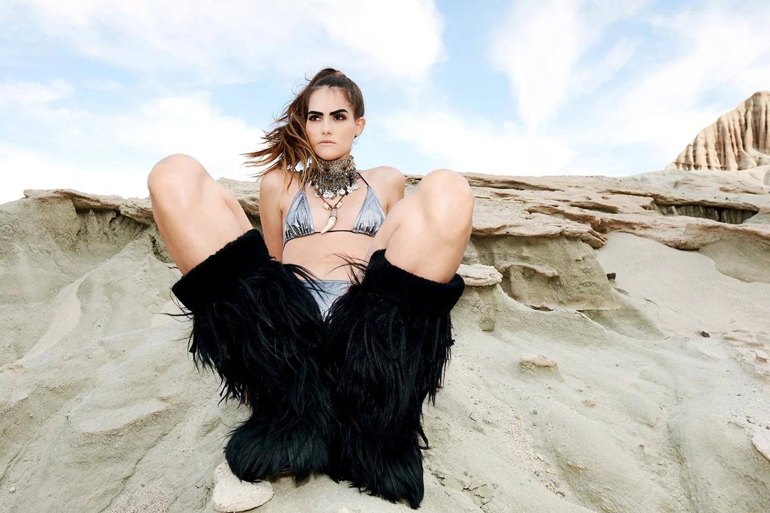 Avant-garde swimwear shot by photographer Daniella Hehmann for Harriet Selling, featuring a model in a patterned bikini and massive black fringe boots seated in a wide-leg pose against a desolate desert horizon.