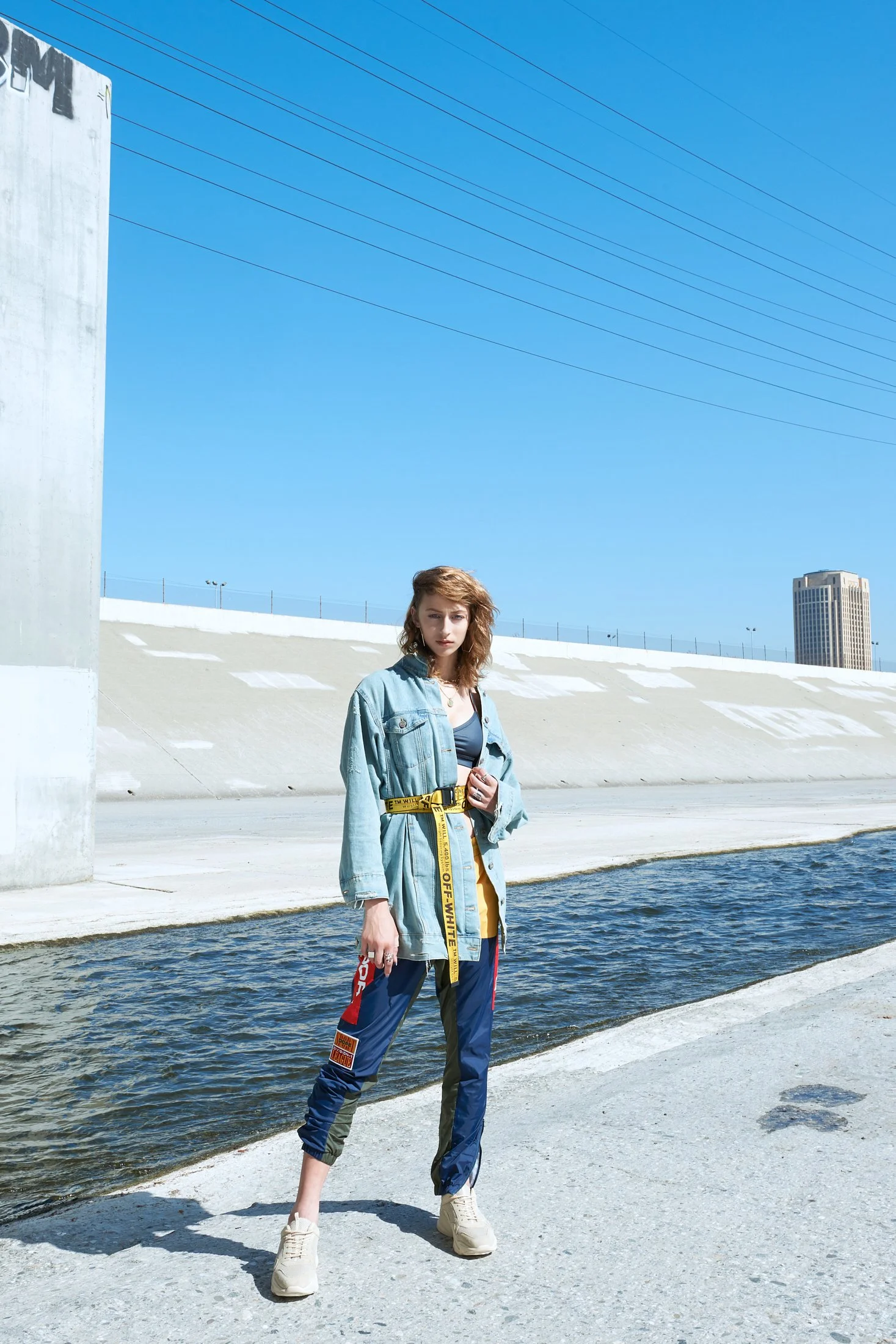 A high-contrast fashion shot by photographer Daniella Hehmann in L.A. featuring a model in a denim jacket with a bold yellow 'Off-White' style belt, captured against the industrial backdrop of the Los Angeles River.