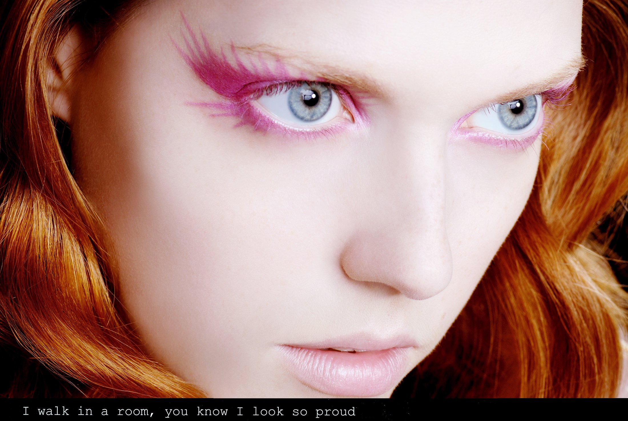 Close-up of a woman with red hair and blue eyes, wearing pink eye makeup with feather-like designs, looking to the side.